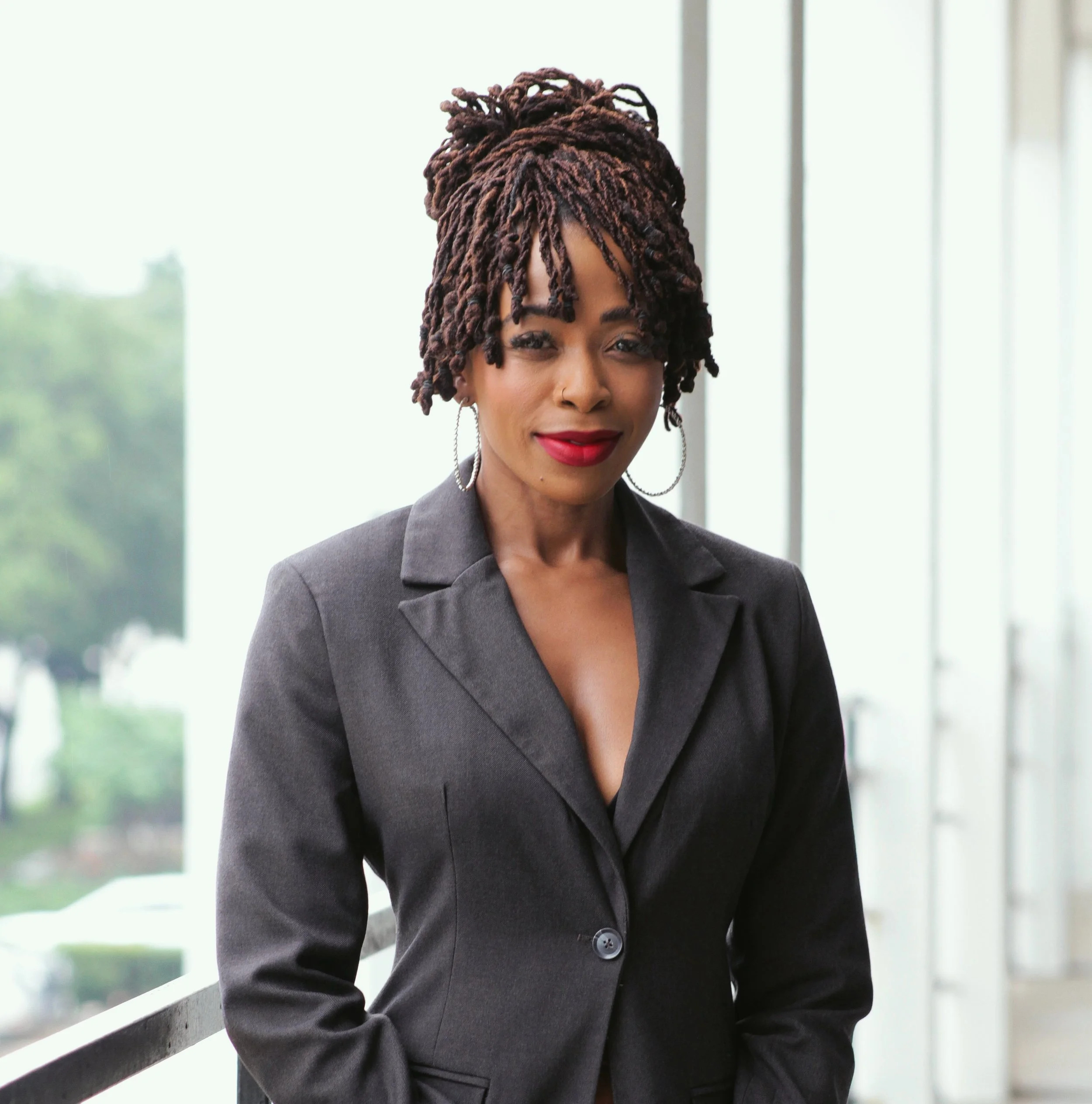 Dr. MaryBeth Asenime is styled brown dreadlocks in a high bun, wearing a gray blazer, standing near a railing in front of a large window.