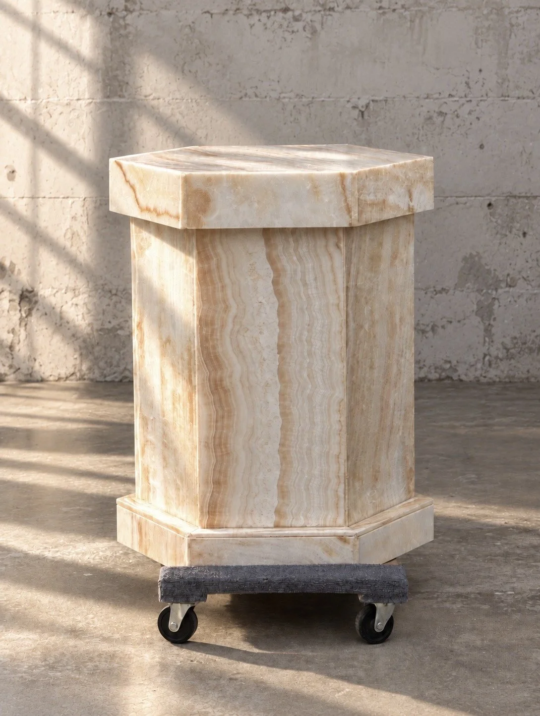 A marble pedestal table on wheels with a beige and cream-colored veined marble surface, placed outdoors against a concrete wall.