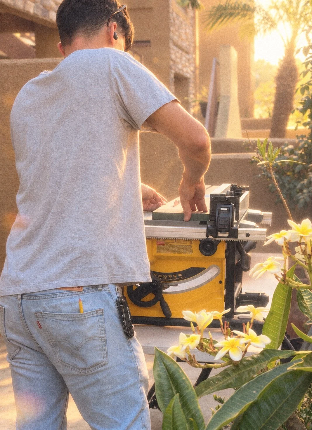 A person working with a table saw outdoors, with yellow flowers and greenery in the foreground, and a warm sunlight background.