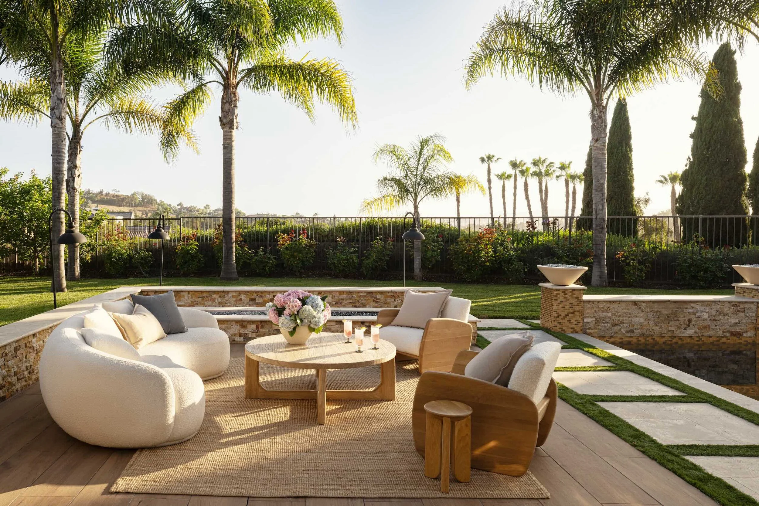 Outdoor patio with curved white furniture, a wooden table with a flower arrangement, surrounded by palm trees and green landscape.