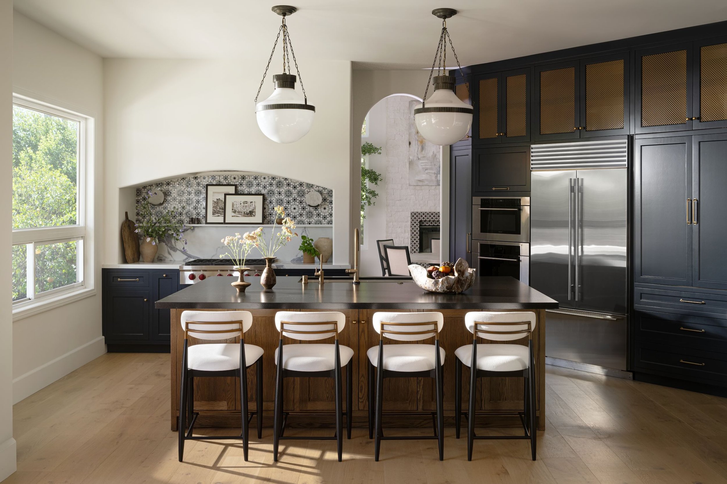 Modern kitchen with navy blue cabinets, a large wooden island with white chairs, pendant lights, and a window showing greenery outside.