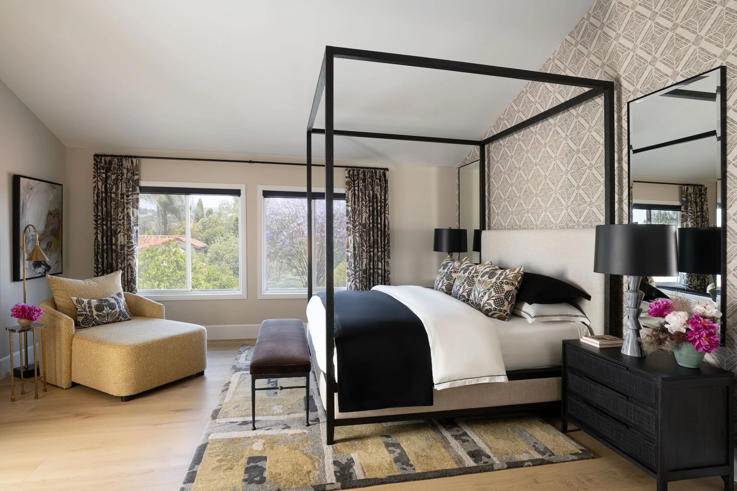 A stylish bedroom with a black four-poster bed, patterned wallpaper, black lamps, and a window with patterned curtains showing a view of trees and houses outside.
