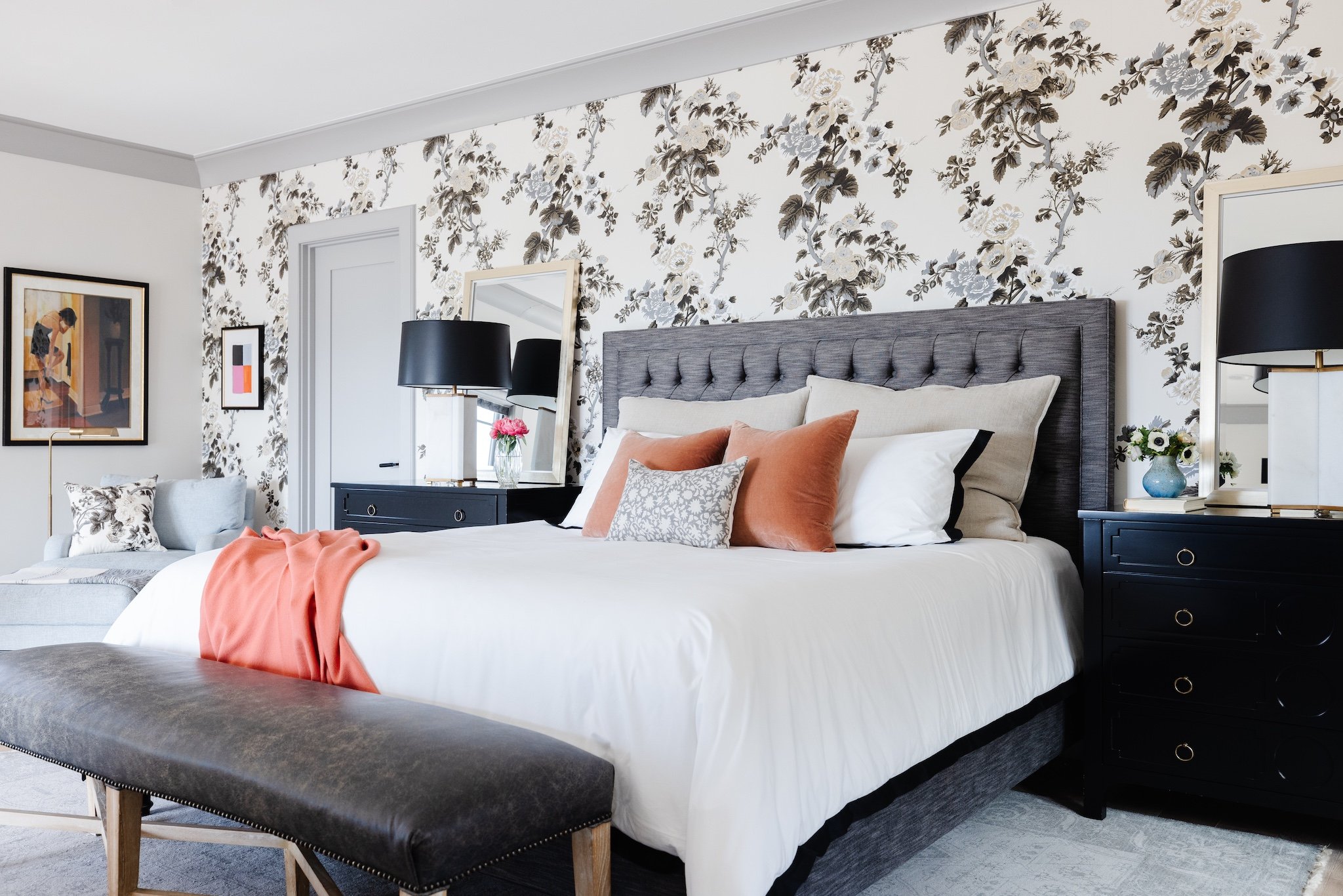 A bedroom with a gray upholstered bed, white and blush pillows, and an orange throw blanket. Behind the bed, there is floral wallpaper with white flowers and darkest gray leaves. On the nightstands, there are black lamps and decorative items like a c