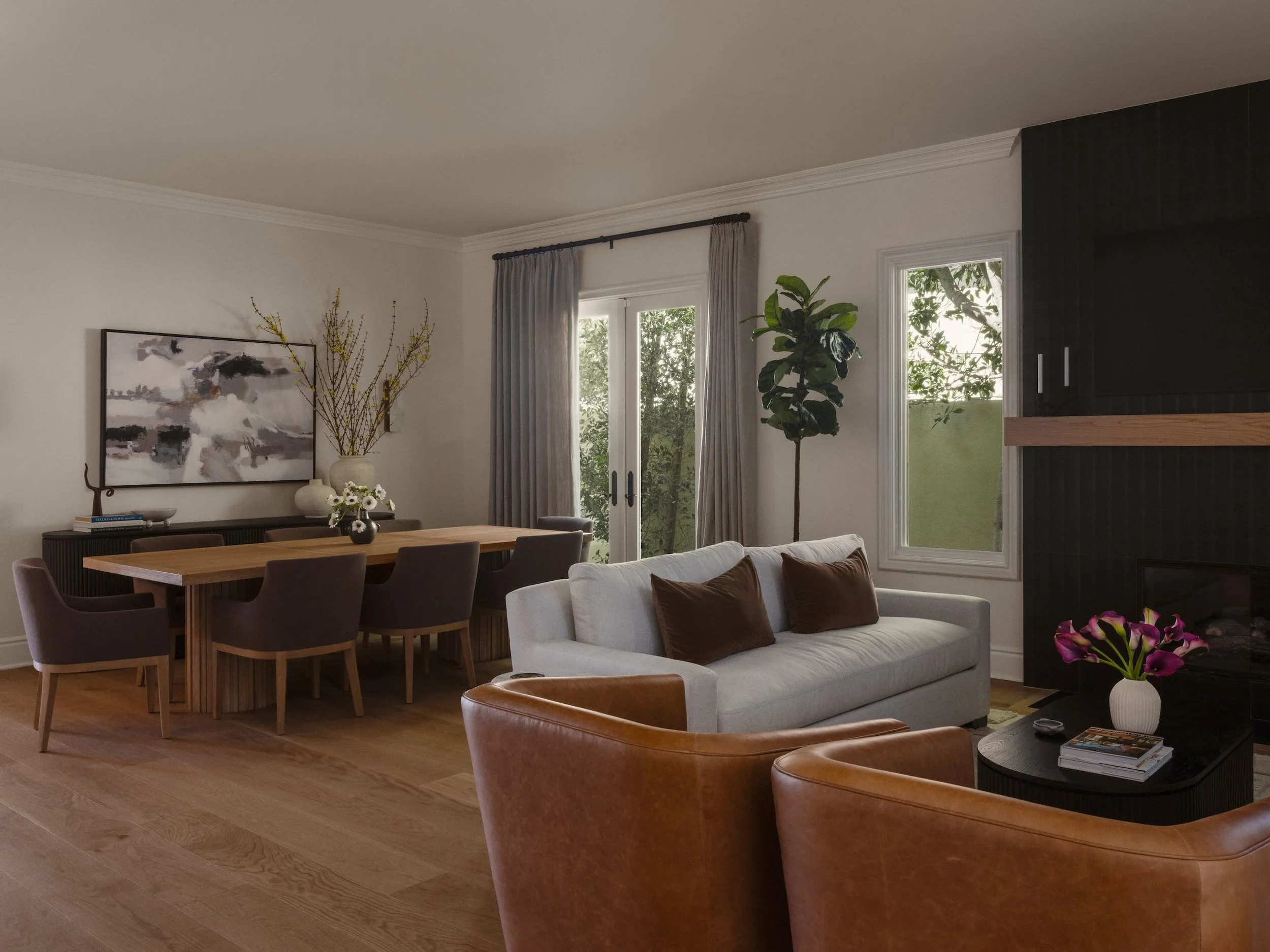 A cozy living room and dining area with large windows, beige curtains, a white sofa with brown pillows, a wooden dining table with dark chairs, and art and plants on the walls.