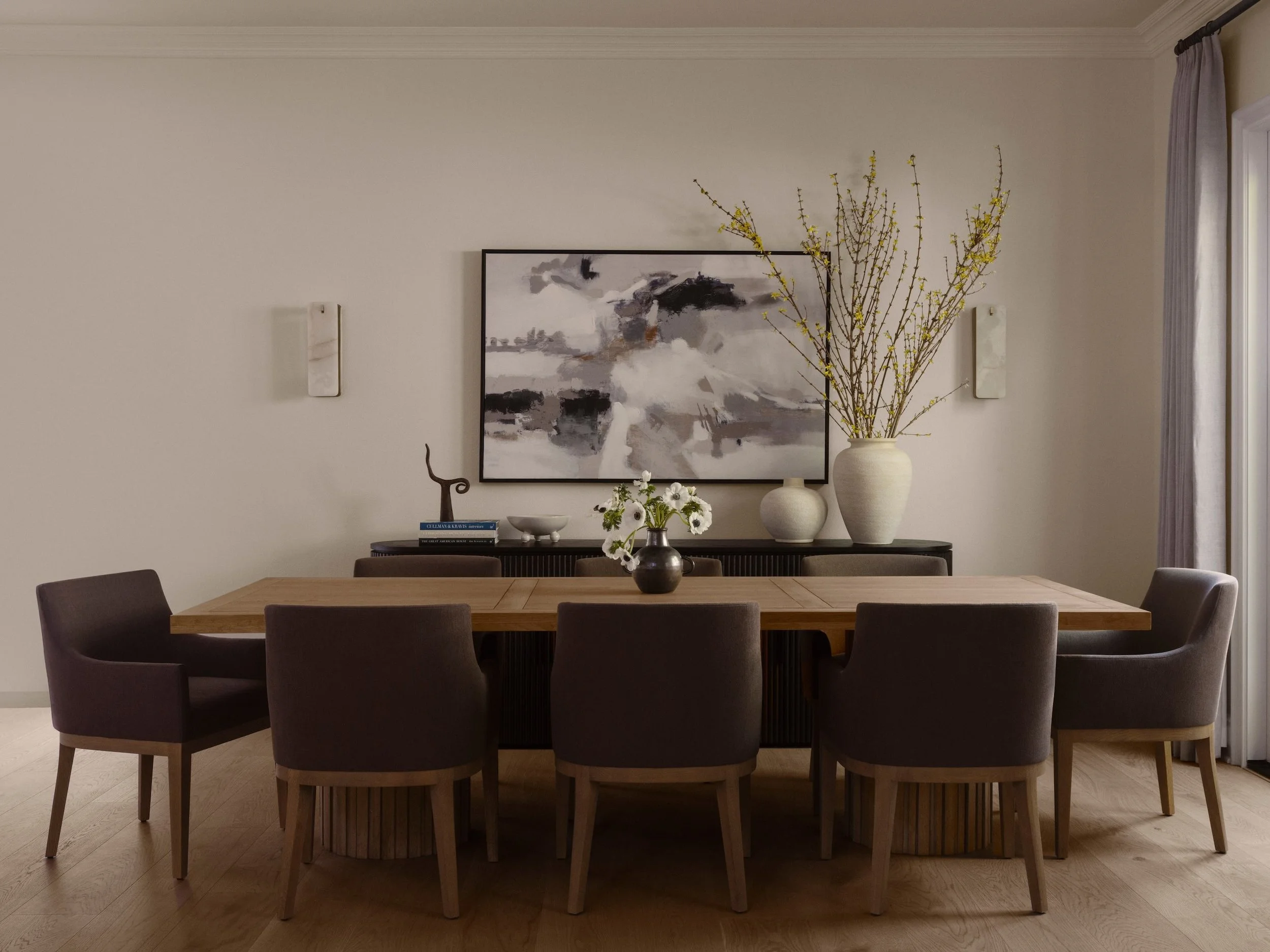 A dining room with a wooden table surrounded by six gray upholstered chairs, a large abstract black and white painting on the wall, and a vase with yellow flowering branches on a sideboard beneath the painting.