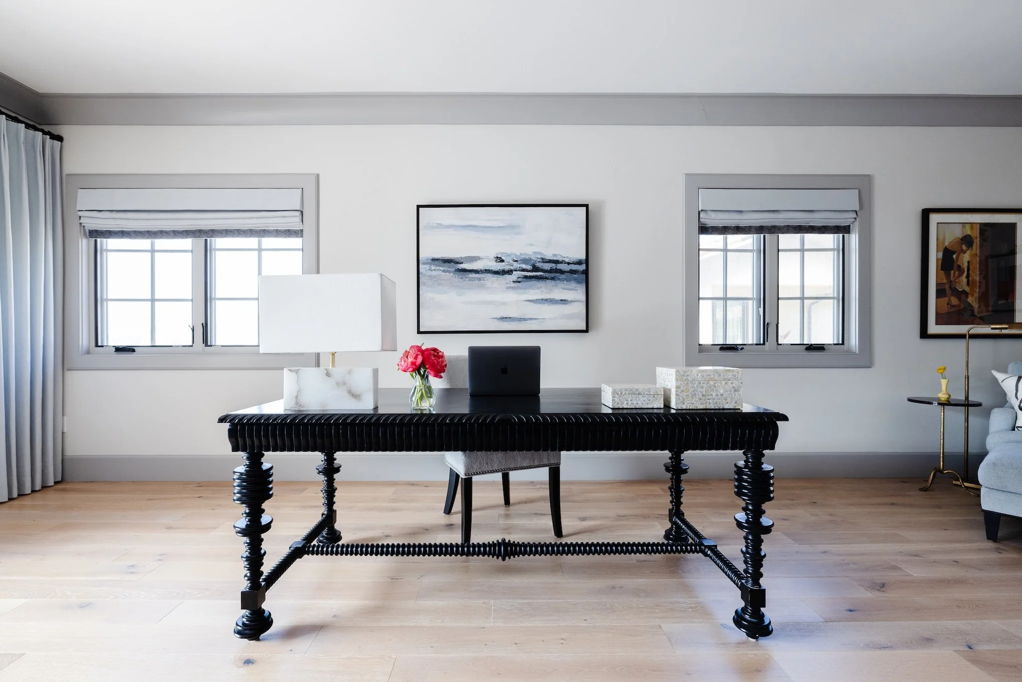 A modern interior room featuring a black table with a twisted wood design, decorated with a white marble lamp, a vase with pink flowers, and decorative boxes. The background includes white walls with framed artwork, two windows with shades, and a gli