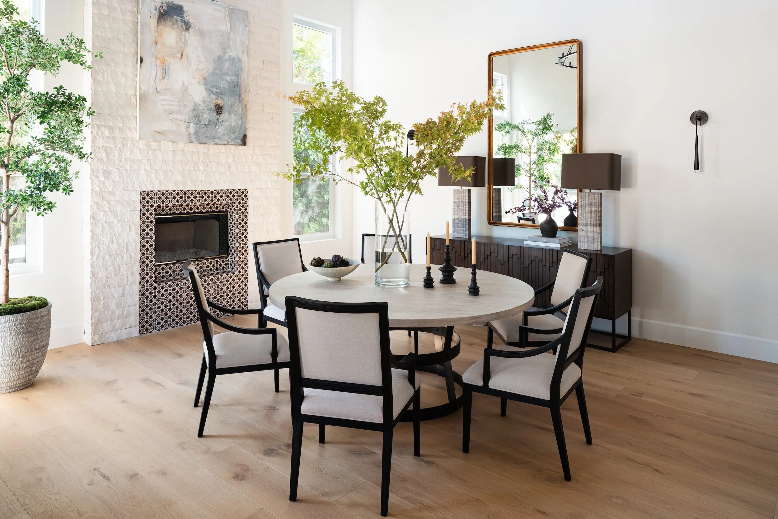Modern dining room with a round table, eight chairs, large windows, potted plants, a white brick fireplace, wall art, mirror, and decorative candles.