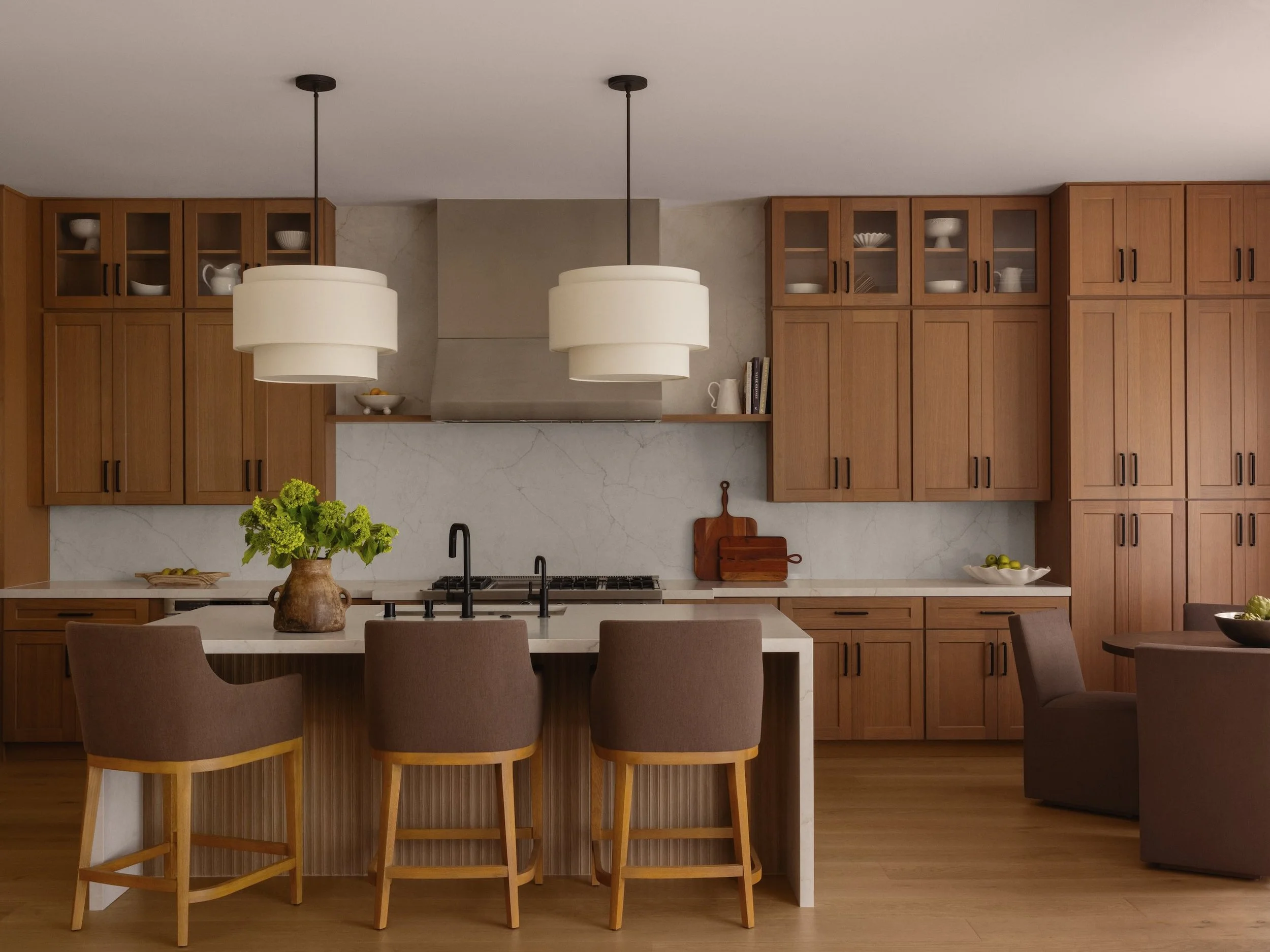 A modern kitchen with wooden cabinets, a white marble countertop, black faucet, two large white pendant lights hanging from the ceiling, a vase of green flowers on the island, and a dining table with beige chairs.