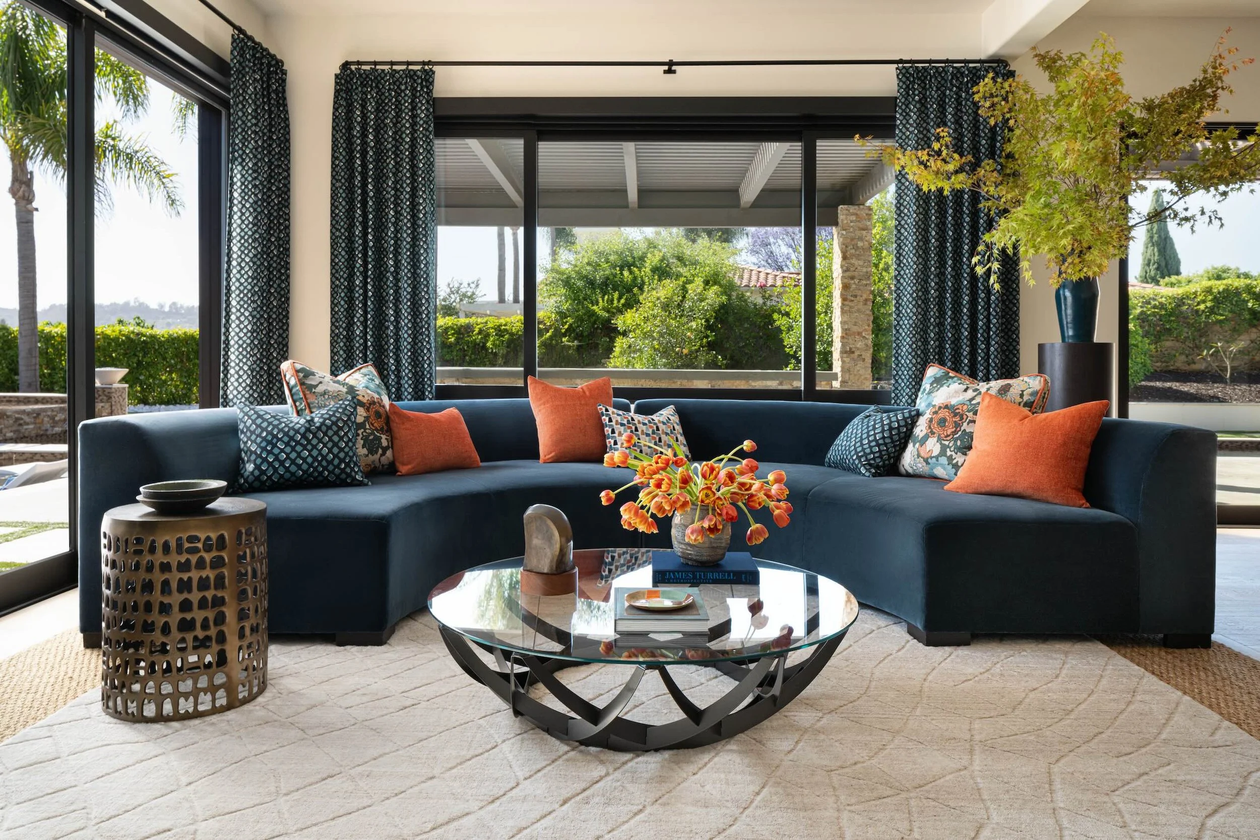 Living room with a large sectional sofa decorated with orange and patterned throw pillows, a round coffee table with a book and a vase of orange flowers, and a sliding glass door leading to an outdoor patio with greenery.