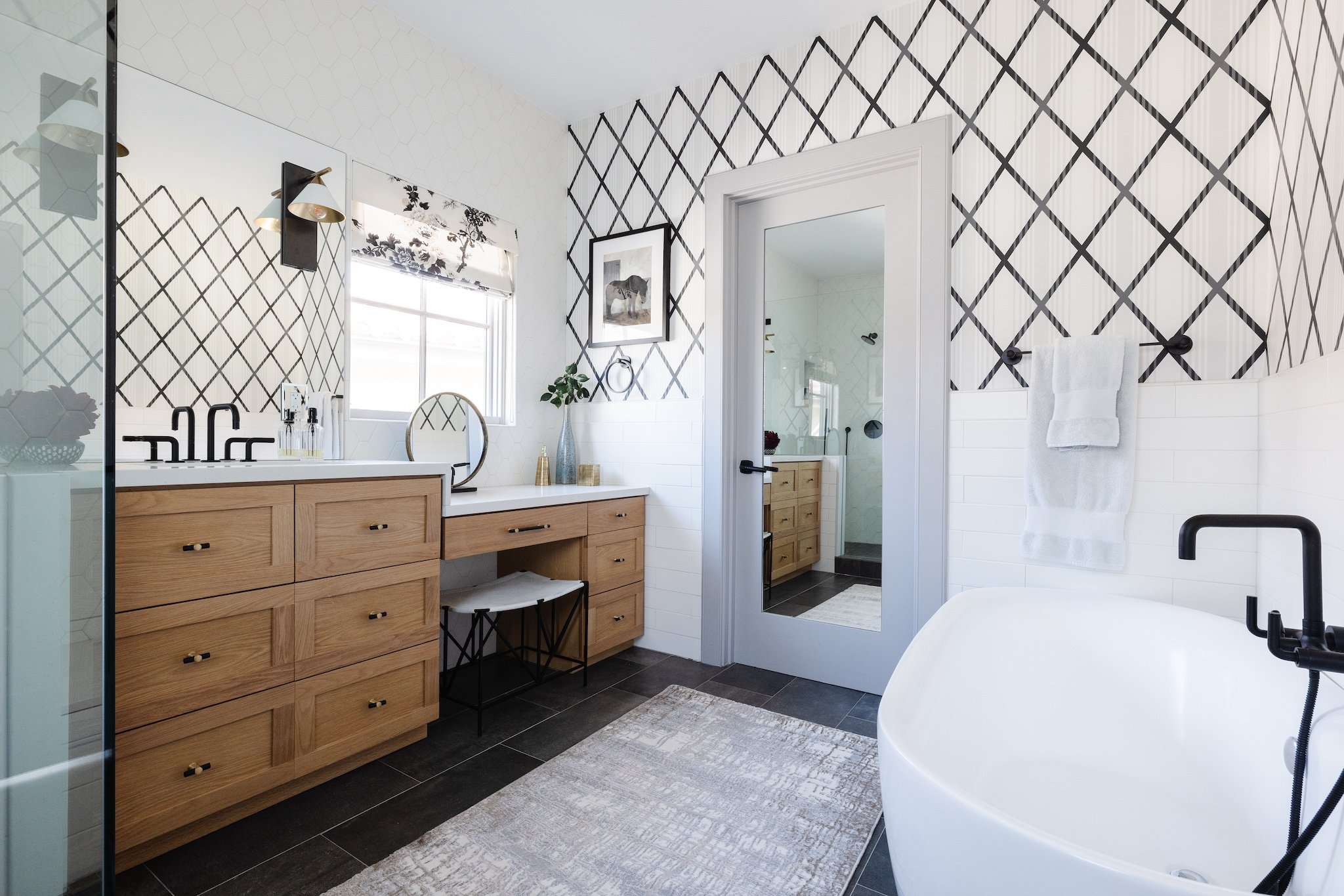 Modern bathroom with black and white geometric wallpaper, a wooden vanity with a white countertop, a large mirror, a framed black and white artwork, and a freestanding bathtub with a black faucet.