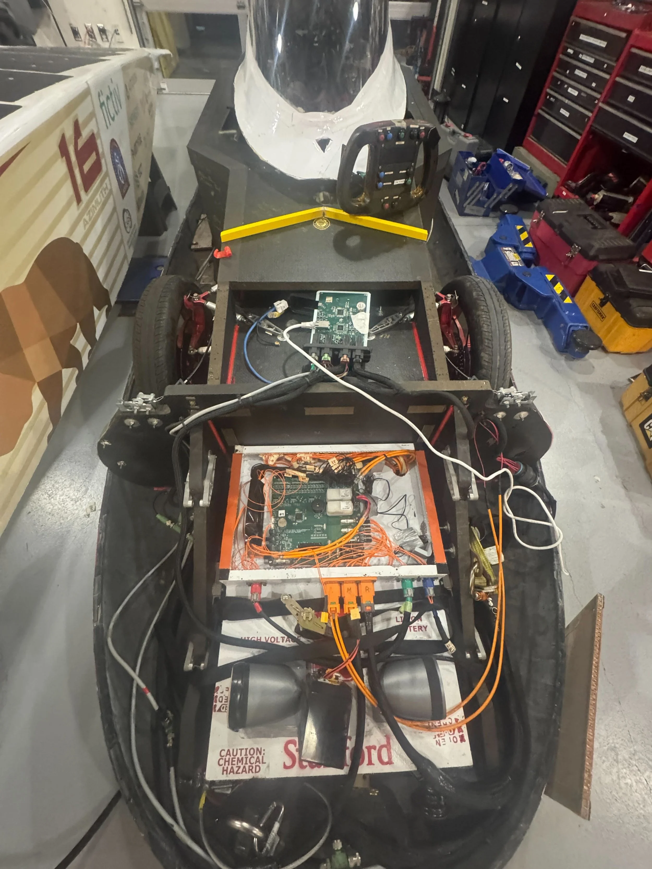 Close-up of an electric race car chassis with electronic components, wiring, and safety labels, inside a garage or workshop setting.