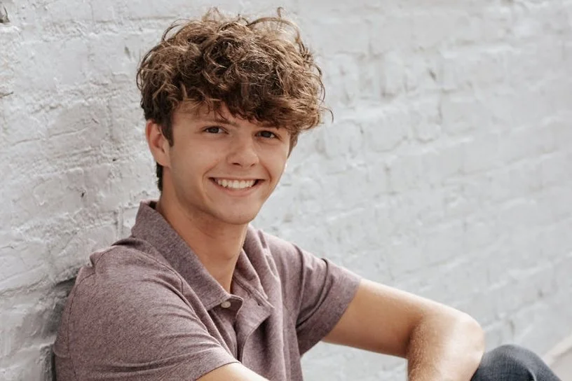 A young man with curly brown hair smiling, sitting against a white brick wall.