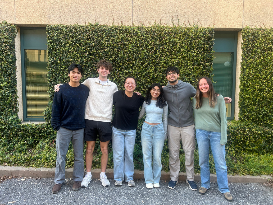 Group of six diverse young adults standing outdoors in front of a leafy hedge, smiling and posing for a photo.