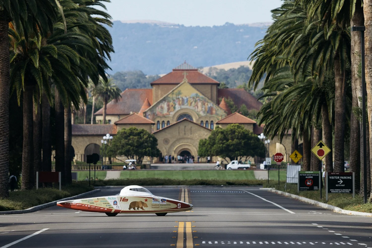 A streamlined, futuristic race car with sponsor logos and a polar bear graphic on its side is parked in the middle of an empty street lined with palm trees. In the background, there is a large historic building with a mural and a red-tiled roof, set against rolling hills.