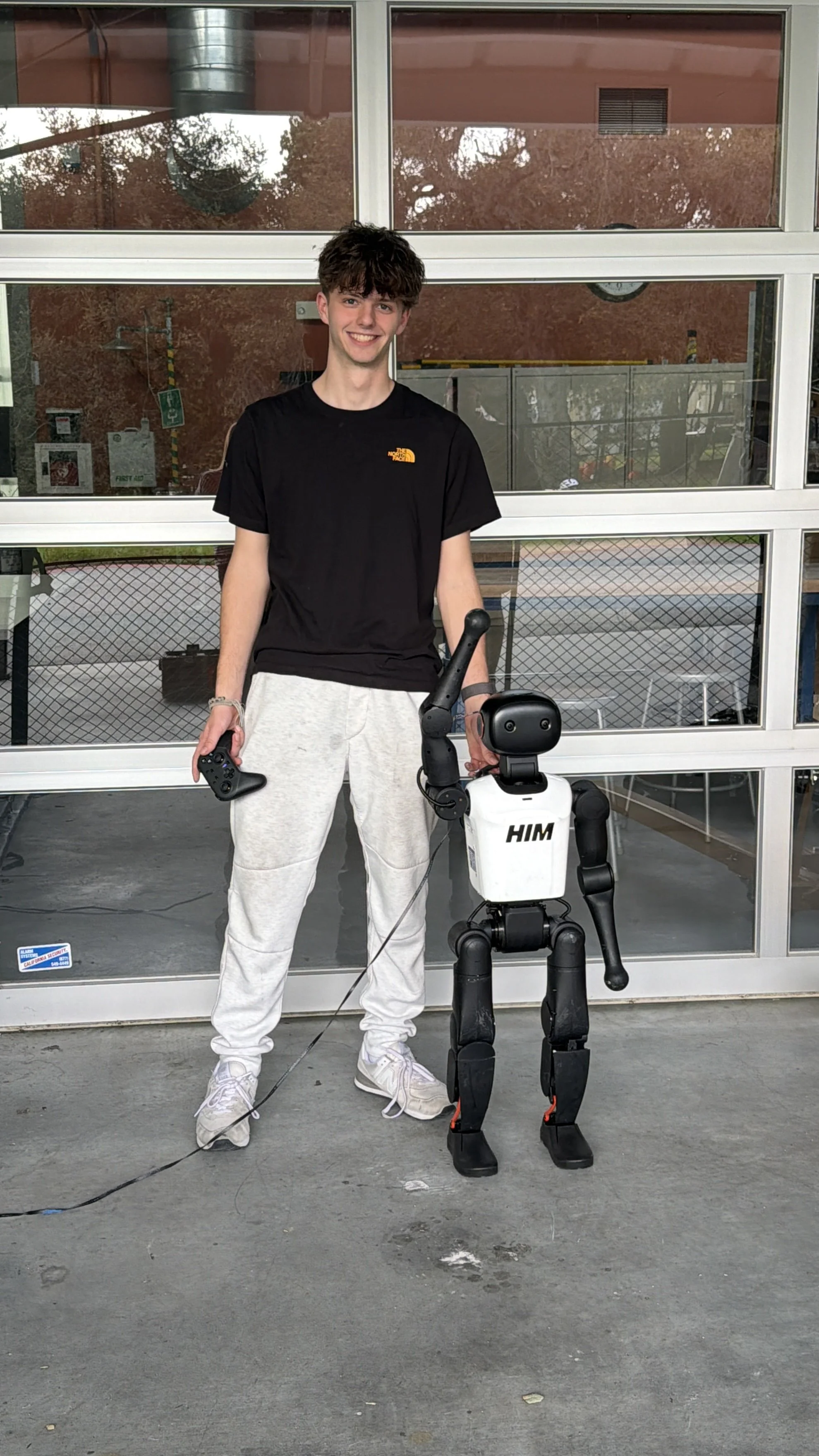 A young man in casual clothing standing outside in front of a glass garage door, holding a remote control, with a robot that has a humanoid appearance and the letters 'HIM' on its chest.