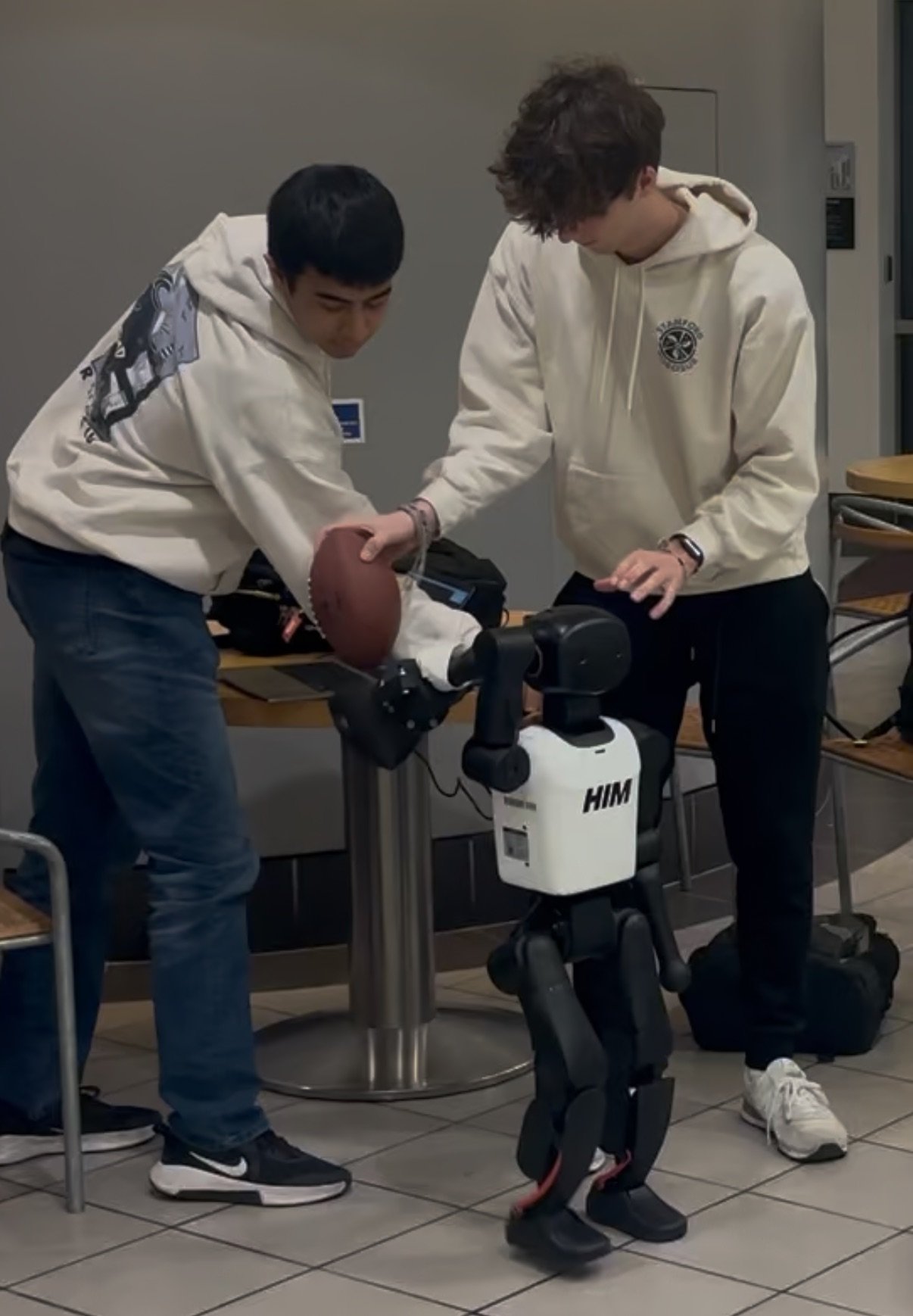 Two young men are working together on a robotic arm, with one holding a basketball. They are in a room with tile flooring, desks, and chairs, wearing casual clothes including hoodies and sneakers.