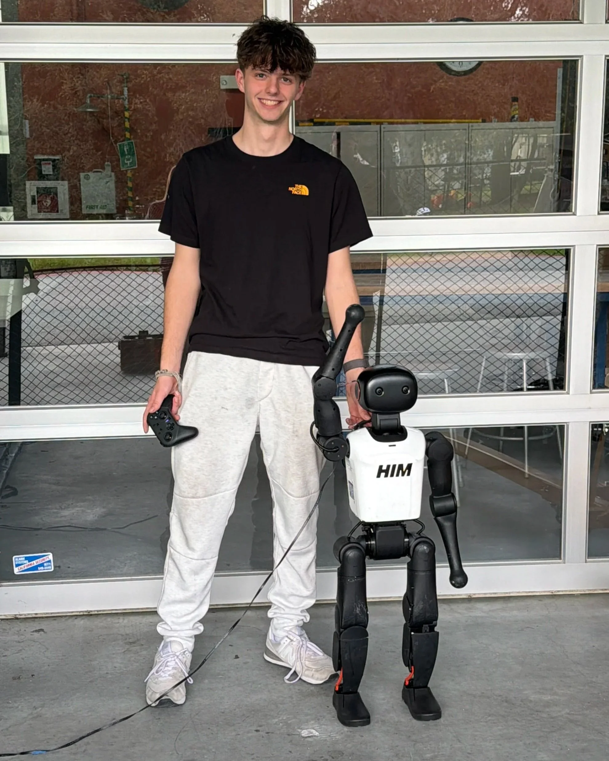 Young man with curly hair smiling, wearing a black North Face T-shirt and white pants, standing outdoors with a robot in front of him, behind glass doors.