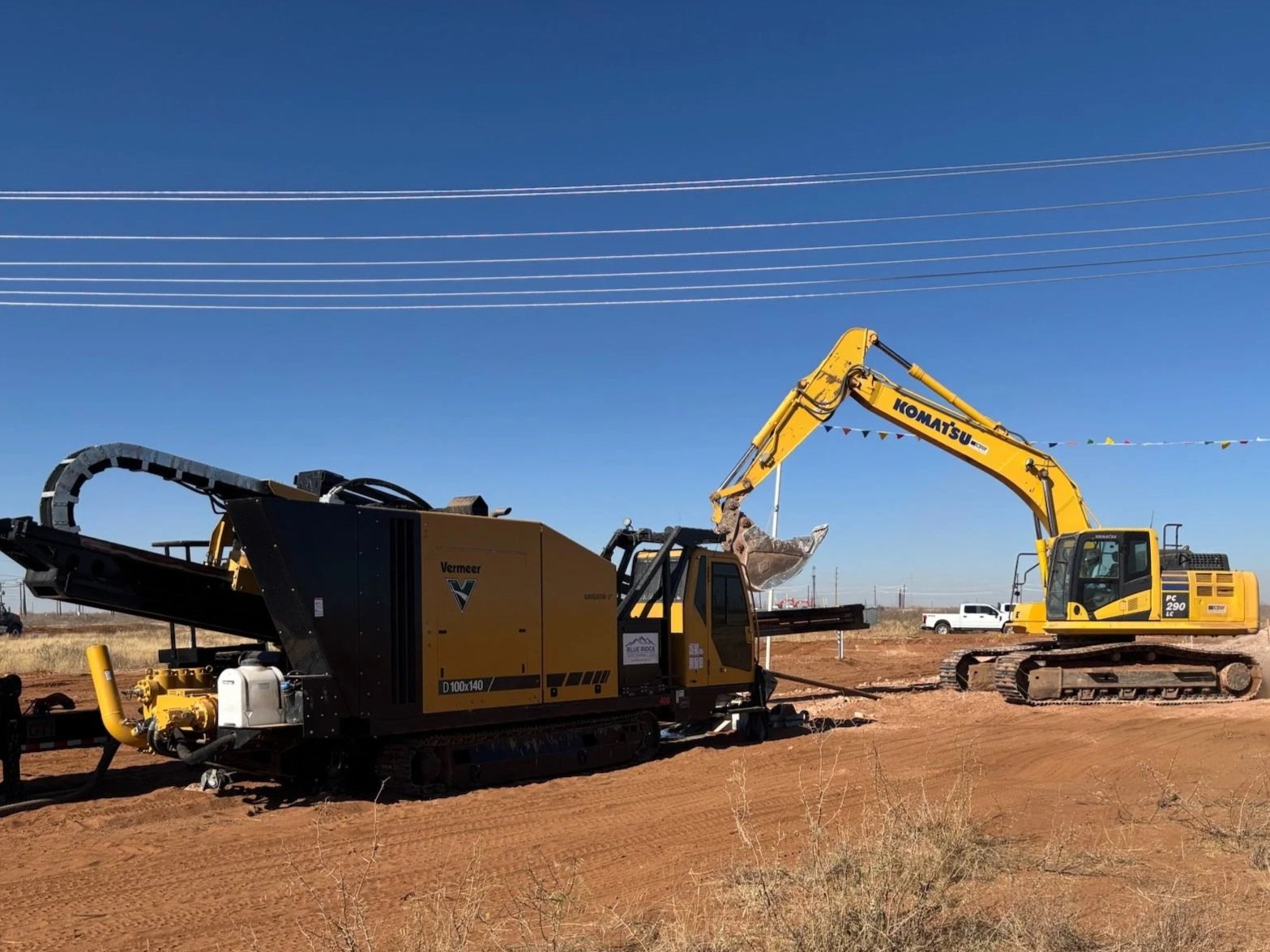 Construction site with a yellow Komatsu excavator and a large specialized Vermeer ground drilling machine on a dirt field under a clear blue sky.