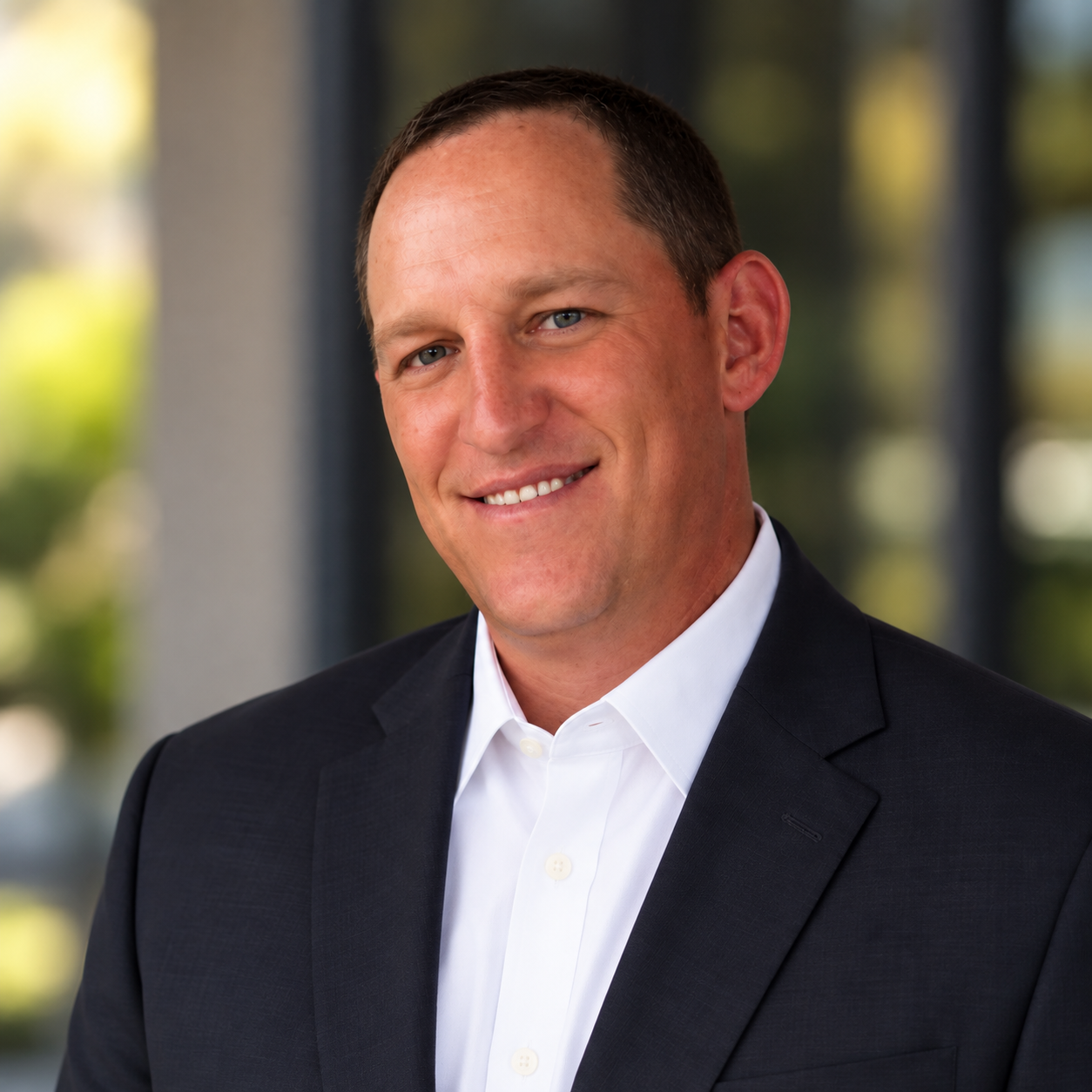 A man in a black suit and white shirt smiling in a professional headshot with a blurred office background.