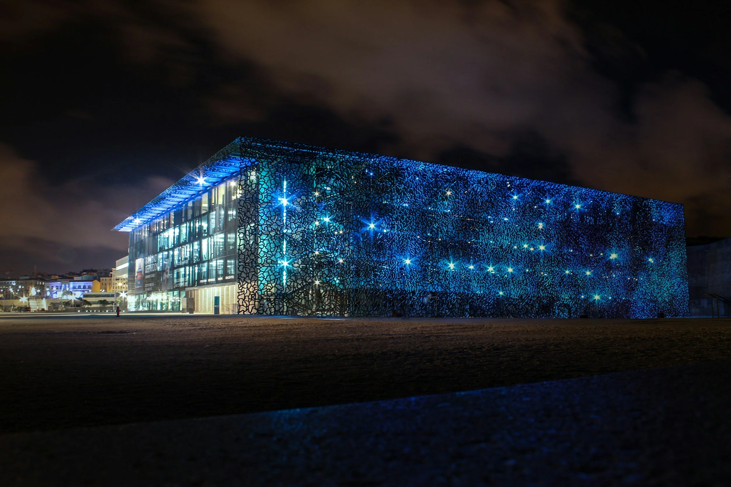 Night view of a modern building with a patterned blue exterior and illuminated windows, under a cloudy sky.