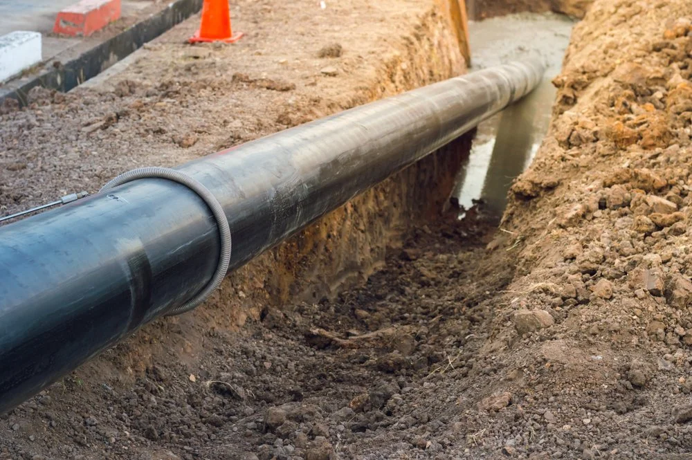 A large black underground pipe installed in a trench at a construction site, surrounded by dirt and rocks, with an orange safety cone in the background.