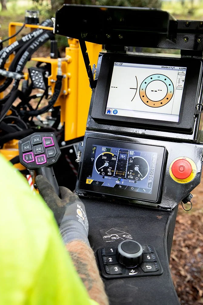 Close-up of a construction worker operating machinery with digital control panels and gauges on a black console, wearing a yellow shirt and gray gloves, with a blurry outdoor background.
