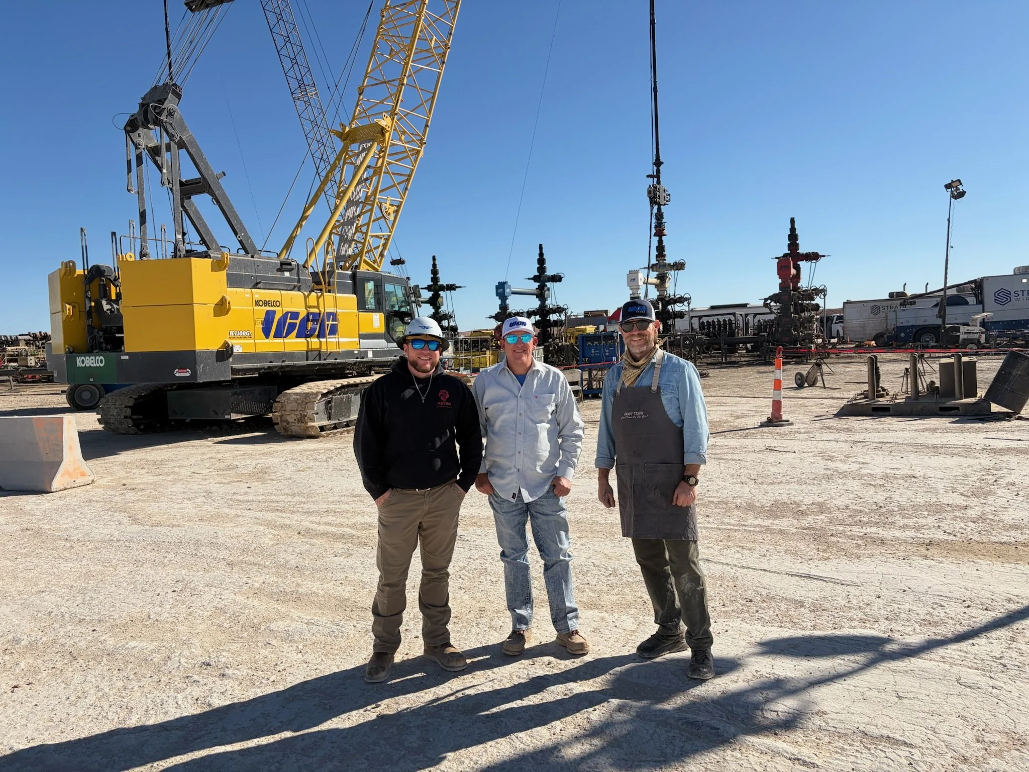 Three men standing outdoors in a construction or industrial site with equipment such as a crane and machinery in the background.