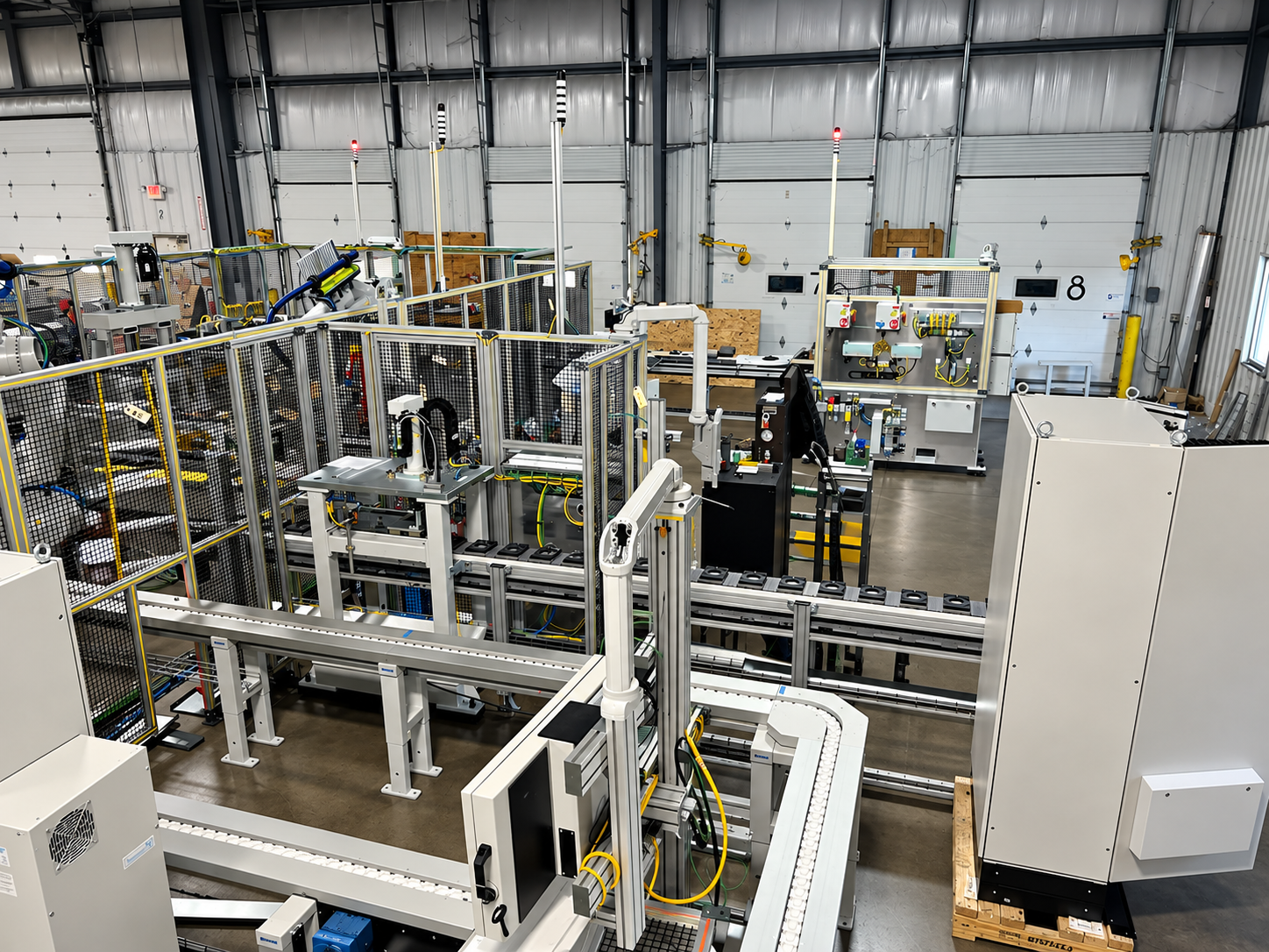 Industrial factory with robotic machinery and equipment, enclosed in safety fencing, inside a warehouse with metal walls and garage doors.