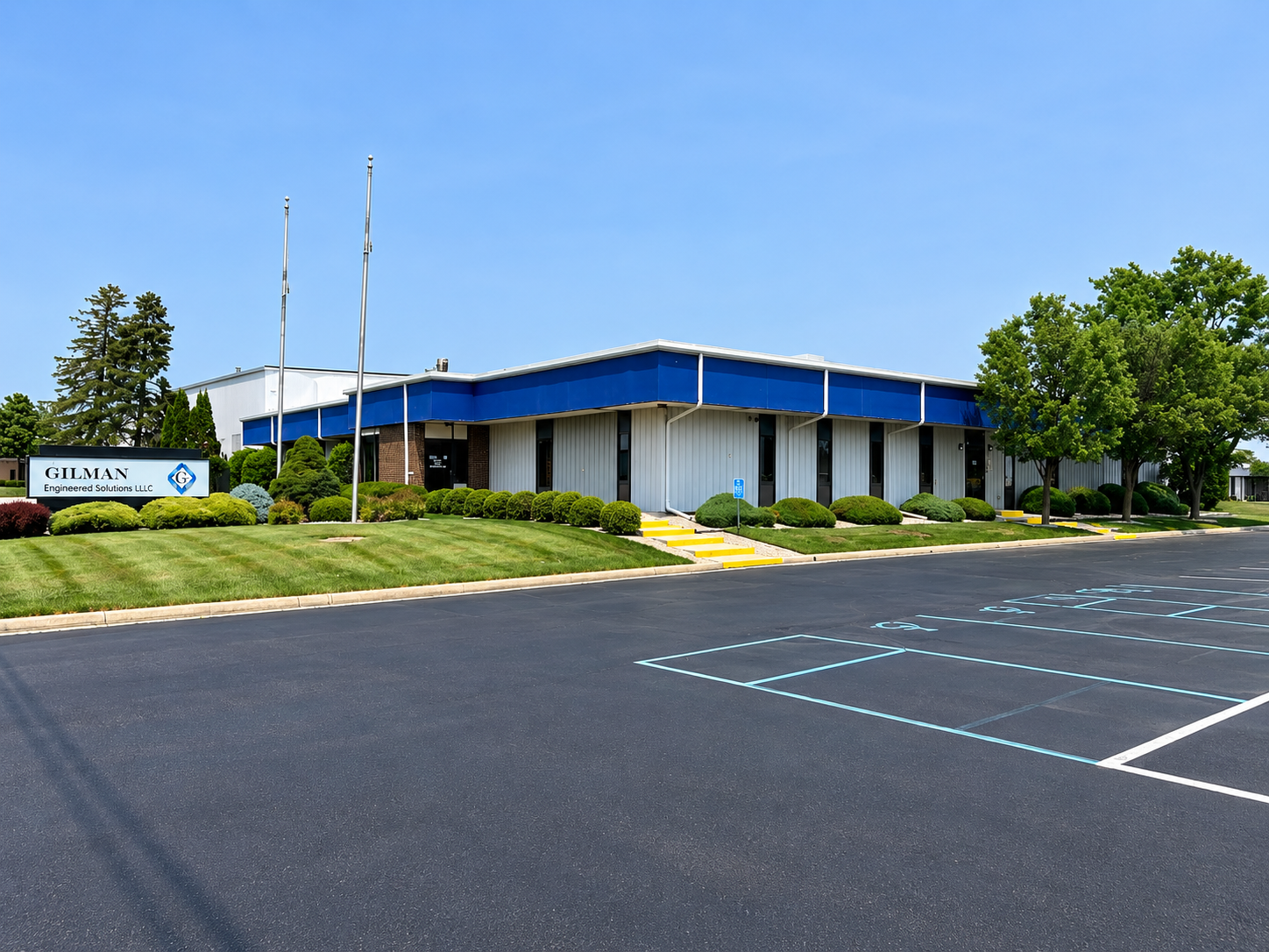 An office building with a sign that reads 'GILMAN Engineered Solutions LLC', surrounded by well-maintained landscaping and a parking lot with designated handicap parking spaces.