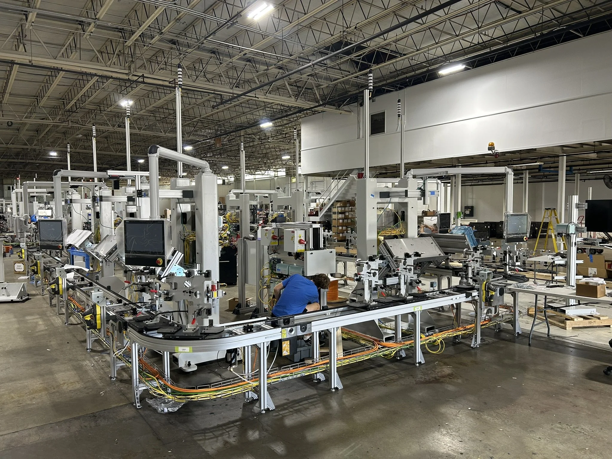 An industrial manufacturing facility with partially assembled machinery, several workers working on equipment, and an exposed ceiling with pipes and lighting.