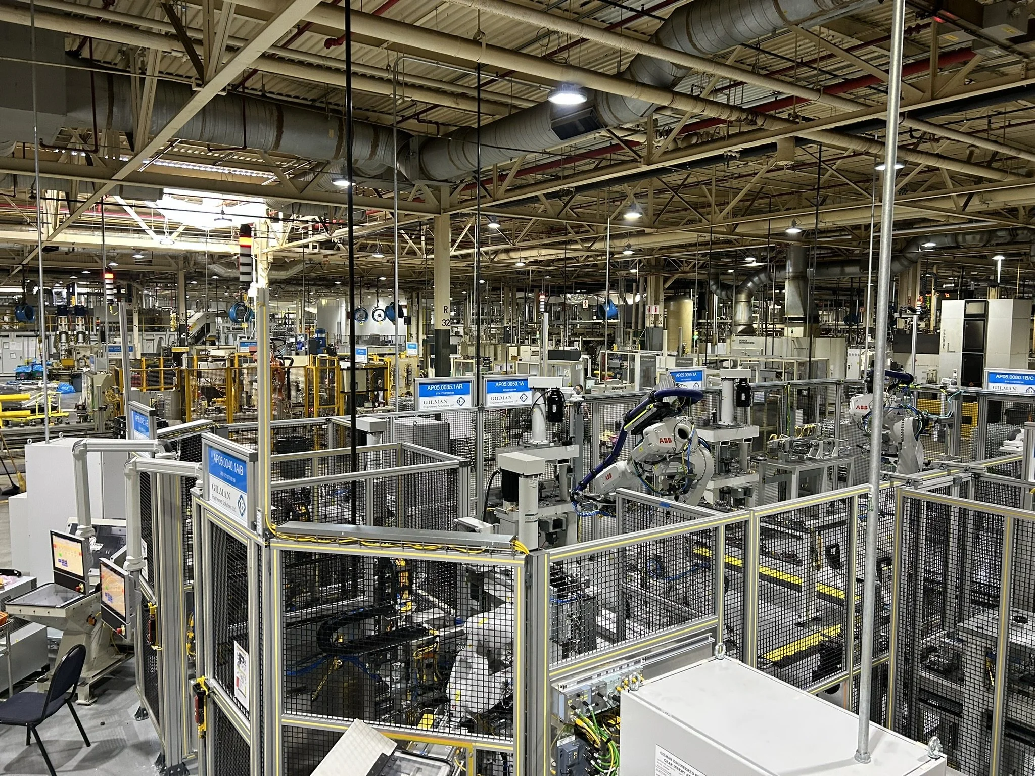 A large industrial manufacturing facility with machinery and equipment, partially enclosed by safety fencing, with a high ceiling featuring ducts and ventilation pipes.