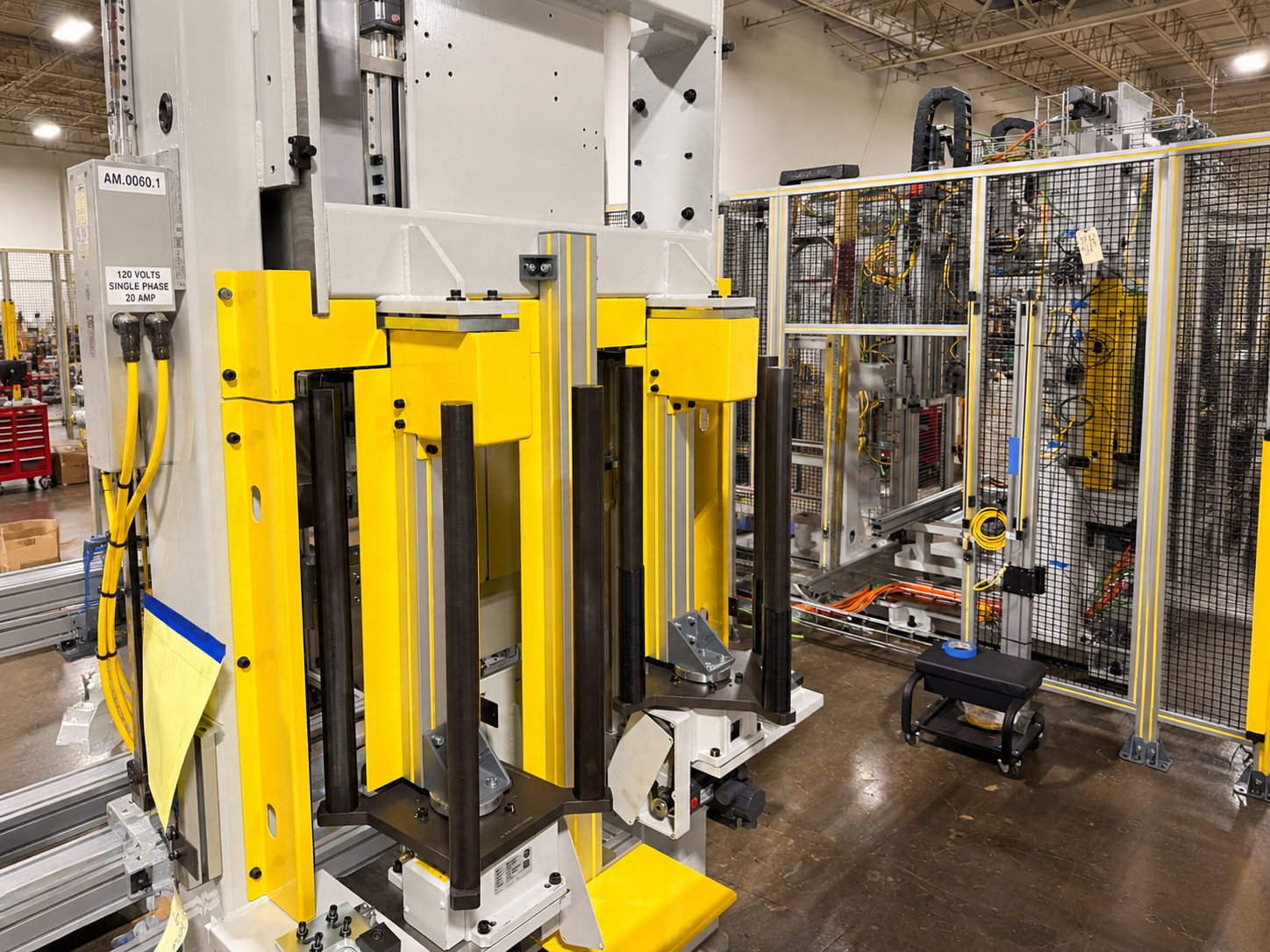 Industrial robotic machinery in a manufacturing plant, with various cables, control panels, and safety fencing, in a spacious warehouse with high ceilings.