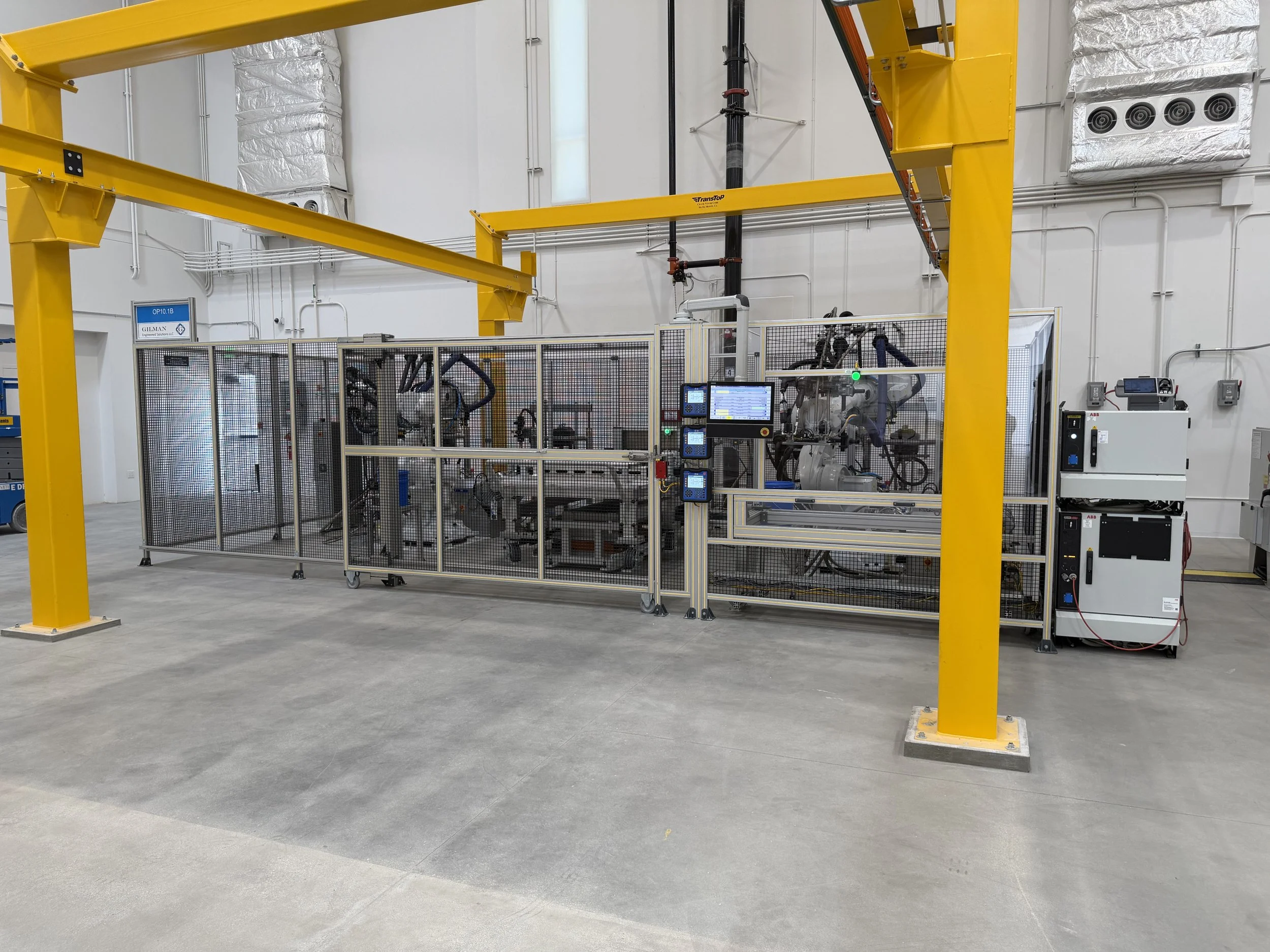 Industrial facility with yellow overhead crane beams, enclosed machinery, control panels and ventilation ducts in a large clean warehouse or factory.