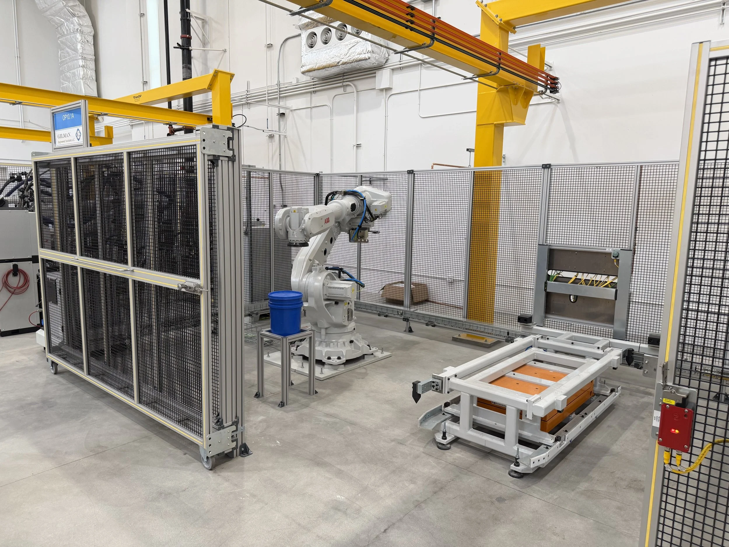 Industrial robotic arm inside safety enclosure in a manufacturing or warehouse facility with overhead yellow safety rails and indoor piping and ventilation.