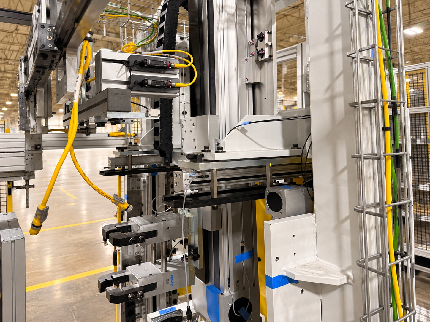 Close-up of industrial manufacturing machinery with metal parts, wires, and yellow hydraulic hoses, situated in a factory setting with a high ceiling.