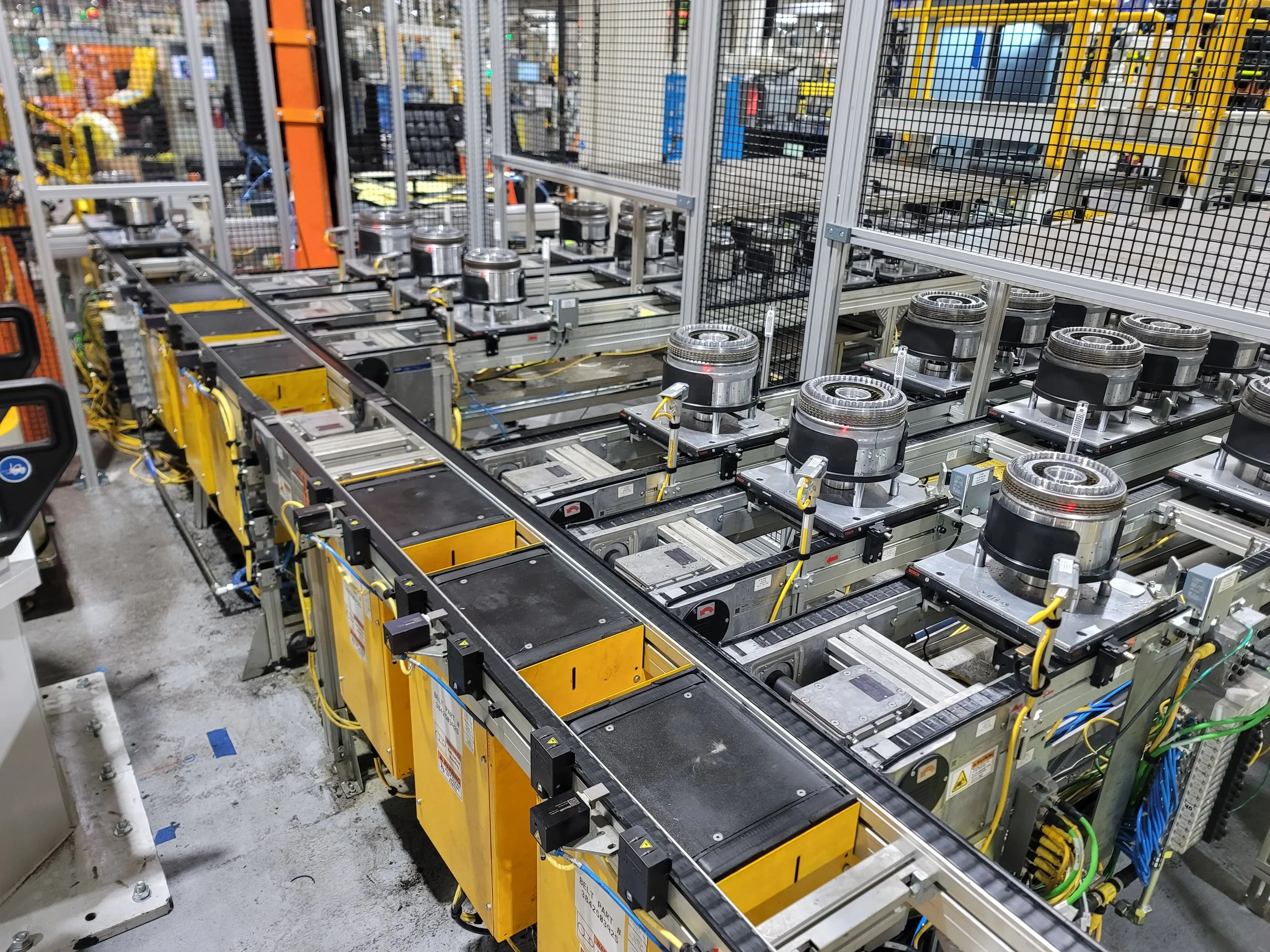 Inside a factory or warehouse, multiple yellow and silver robotic machines and conveyor belts, surrounded by a metal mesh fence, are visible.