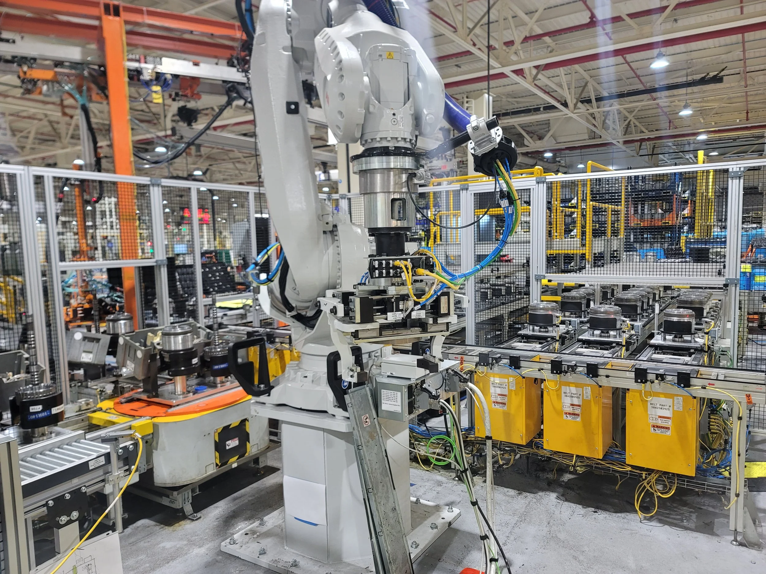 An industrial robotic arm with a white and gray body working inside a manufacturing plant, surrounded by yellow and black safety barriers, with various machinery and overhead lights in the background.