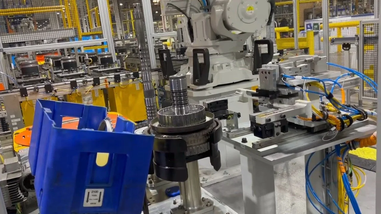 Industrial manufacturing setting with robotic machinery and components, including a large gray robotic arm, metal gears, and various tools and wiring on a worktable.