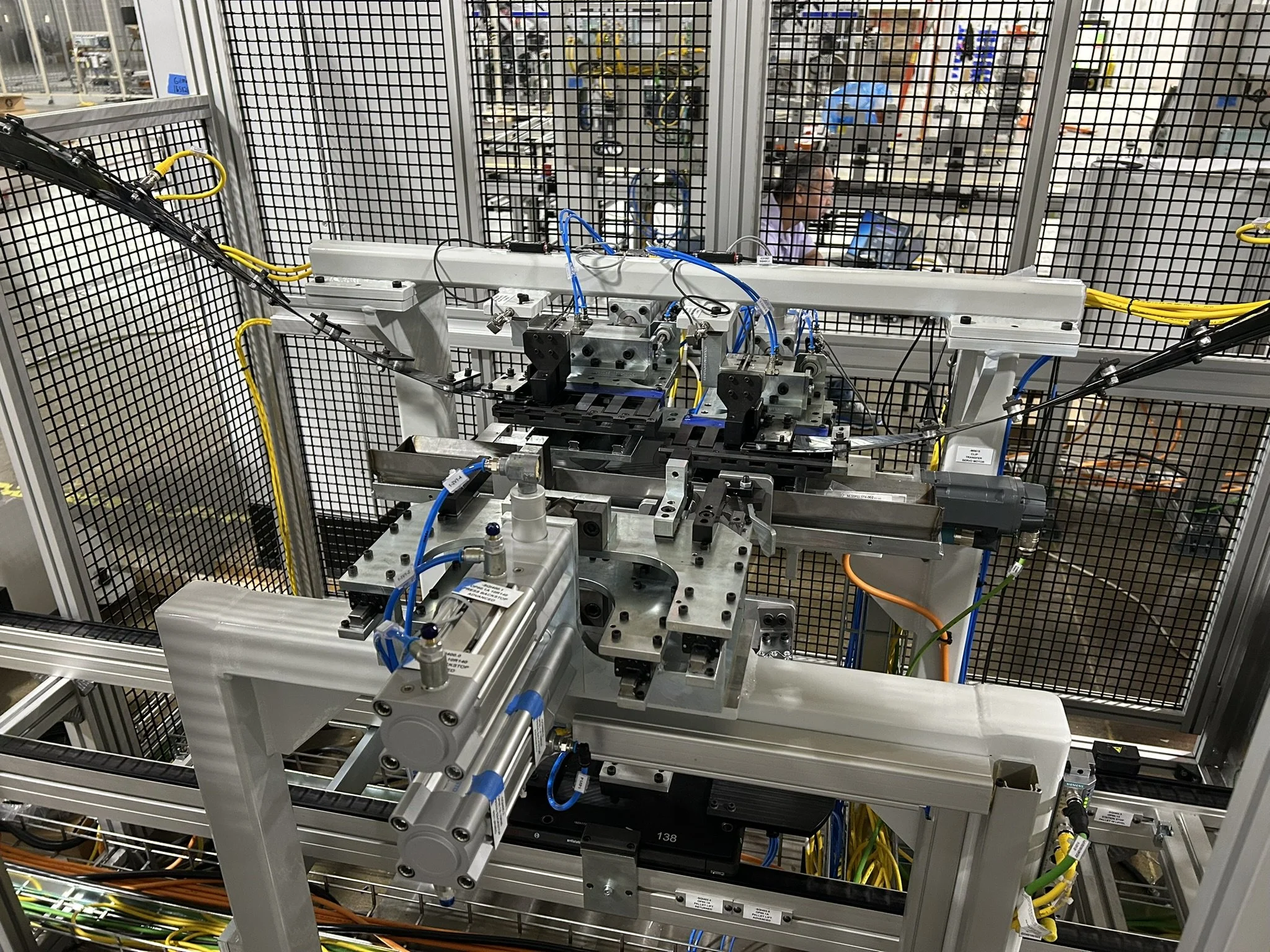 Close-up of industrial machinery with wires and mechanical parts enclosed by metal safety fencing in a manufacturing or laboratory setting.