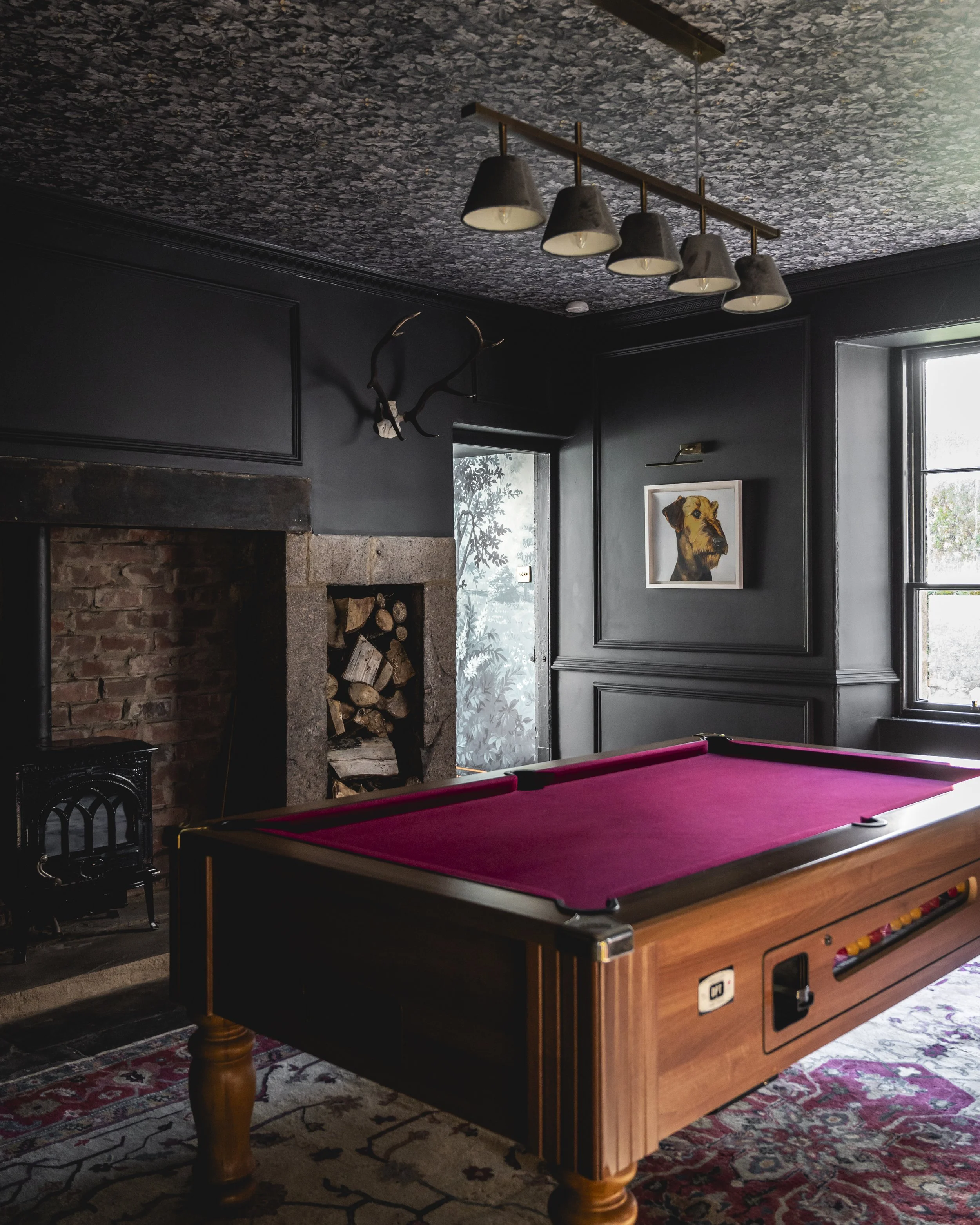 A room with a wood pool table with a red felt surface, a brick fireplace with stacked firewood, a painting of a dog, a chandelier with multiple downward-facing lights, a window, and a wall with black paneling and a deer antler decoration.