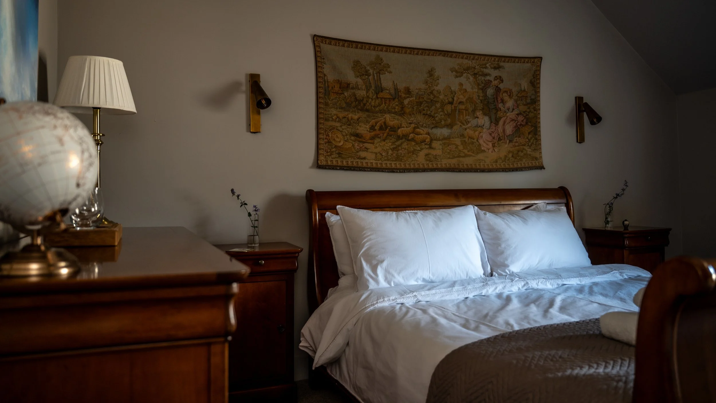 A cozy bedroom featuring a wooden bed with white linens, two nightstands with small vases and flowers, a wall tapestry depicting a pastoral scene, and wall-mounted reading lamps.