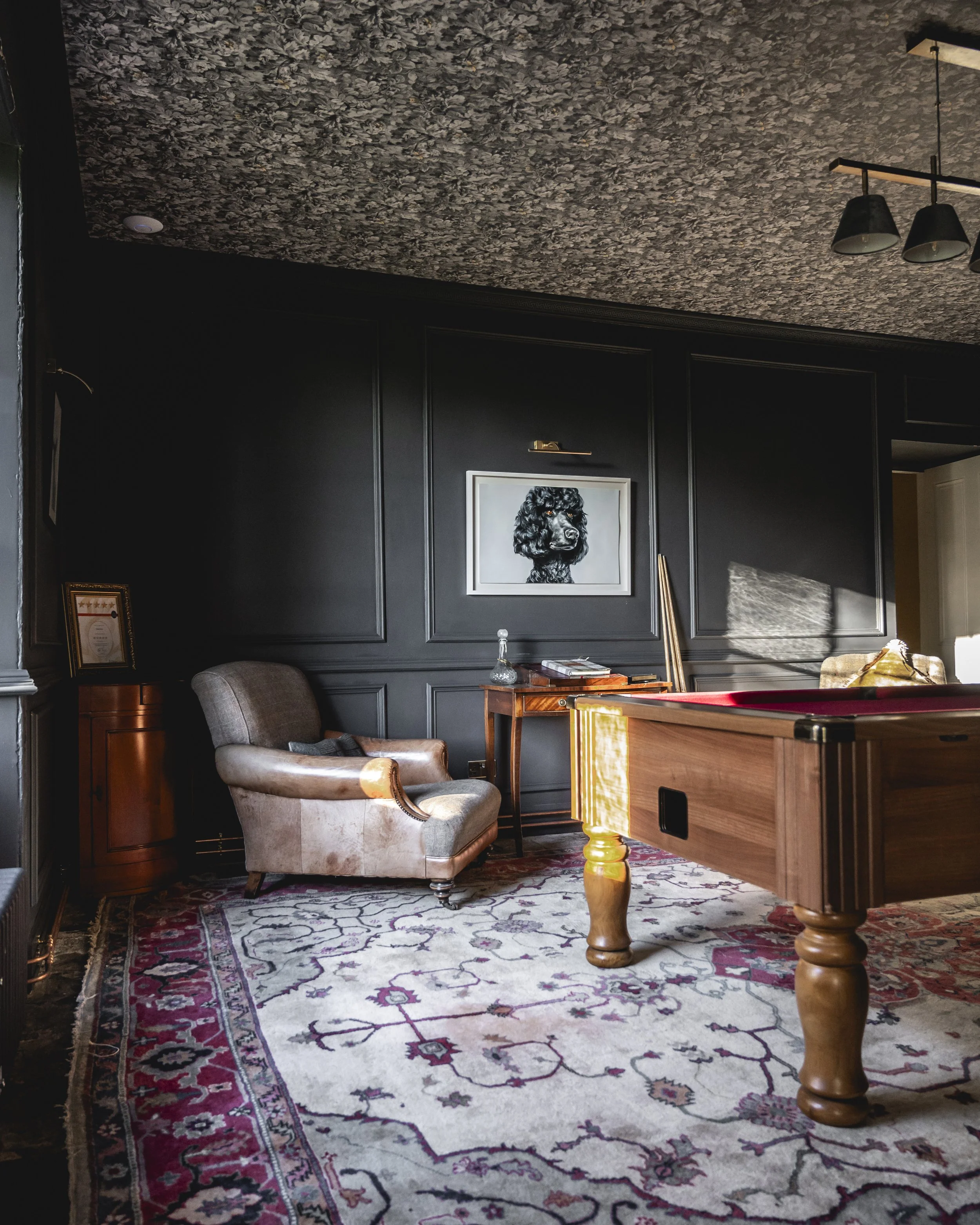 An interior room with dark wall paneling, a patterned carpet, and eclectic furniture including an armchair, a small table with a glass bottle, and a pool table. There is a framed black dog portrait on the wall and modern light fixtures overhead.