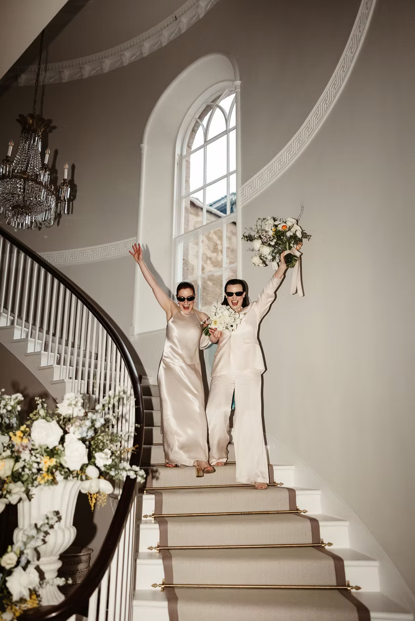 Two women, wearing white outfits and sunglasses, celebrating on a staircase with floral bouquets, in a bright, elegant interior with large arched window and chandelier.