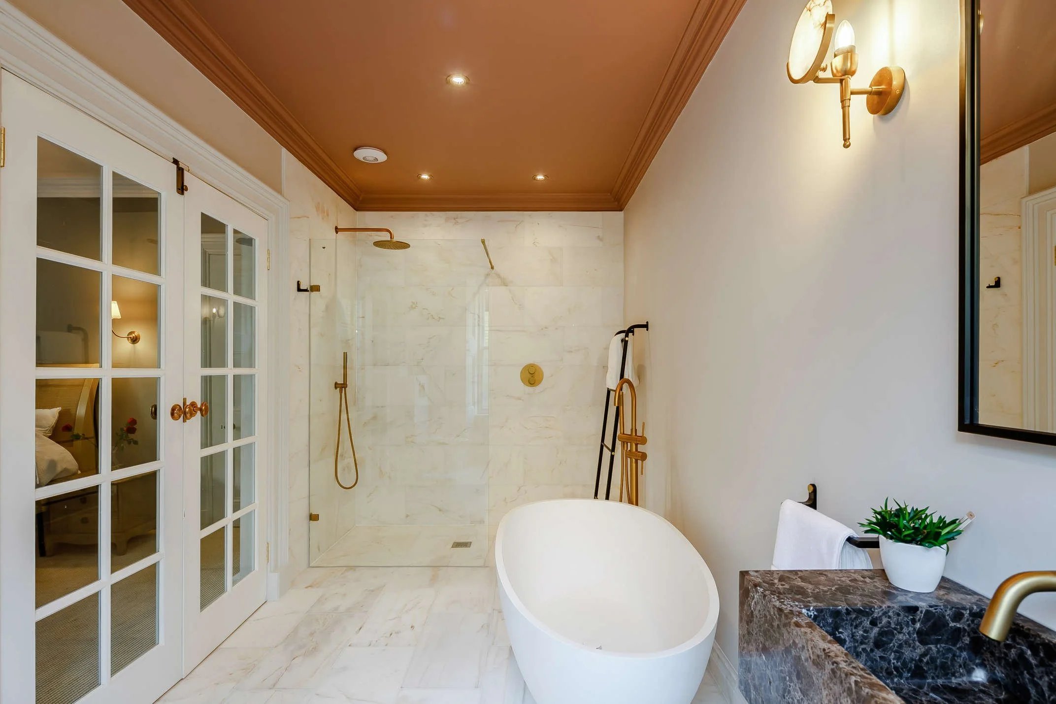 Modern bathroom with a freestanding bathtub, marble shower area with gold fixtures, white walls, a wall-mounted mirror, a marble countertop with a potted plant, and white French doors leading to a bedroom.
