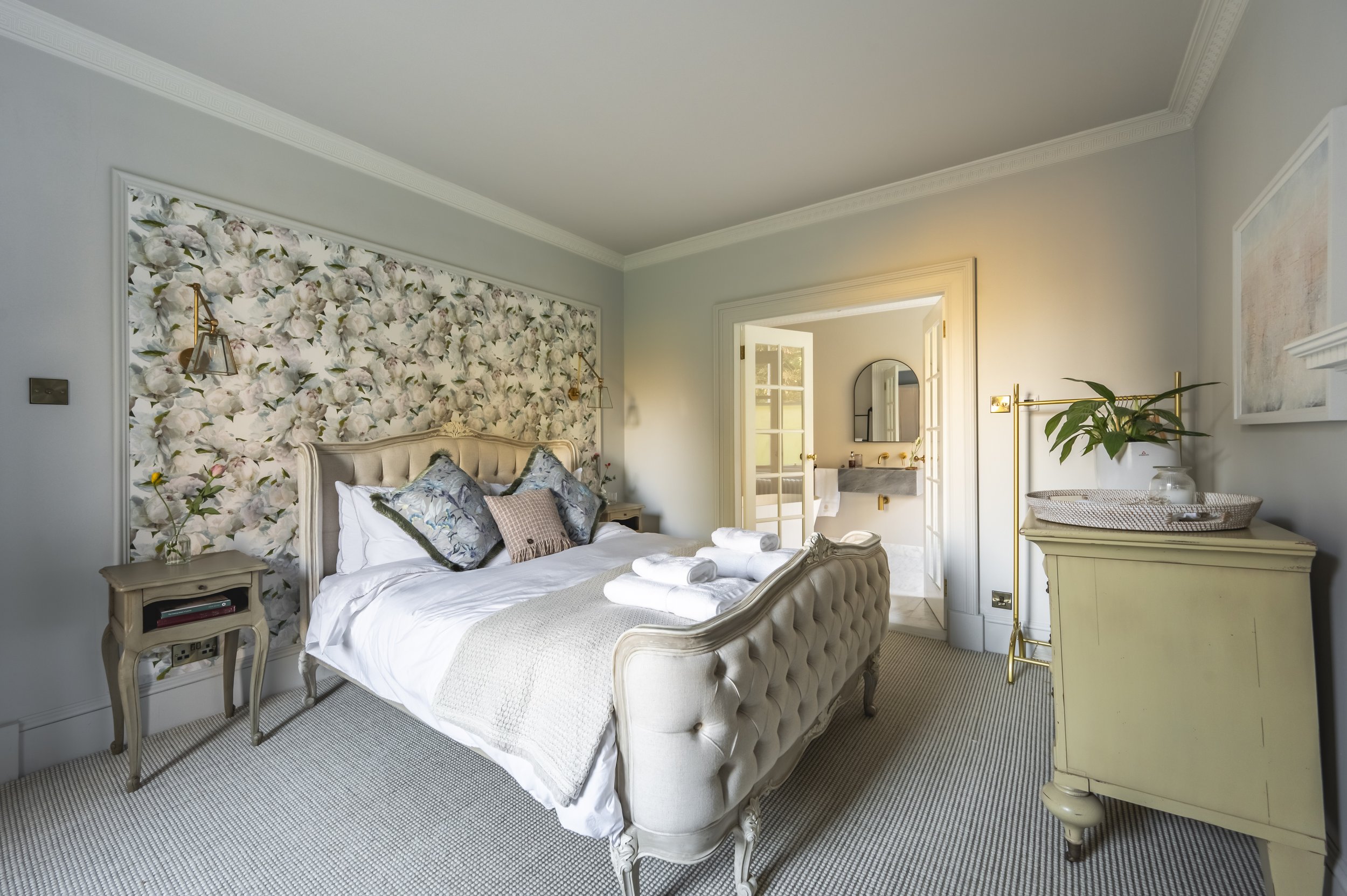A luxurious bedroom with a cream tufted bed and floral wallpaper behind the headboard. The bed has decorative pillows and neatly rolled towels. There are pale yellow nightstands, gold wall sconces, a wicker basket with a plant, and a dresser. A view into an adjacent bathroom with a granite sink is visible through a doorway.