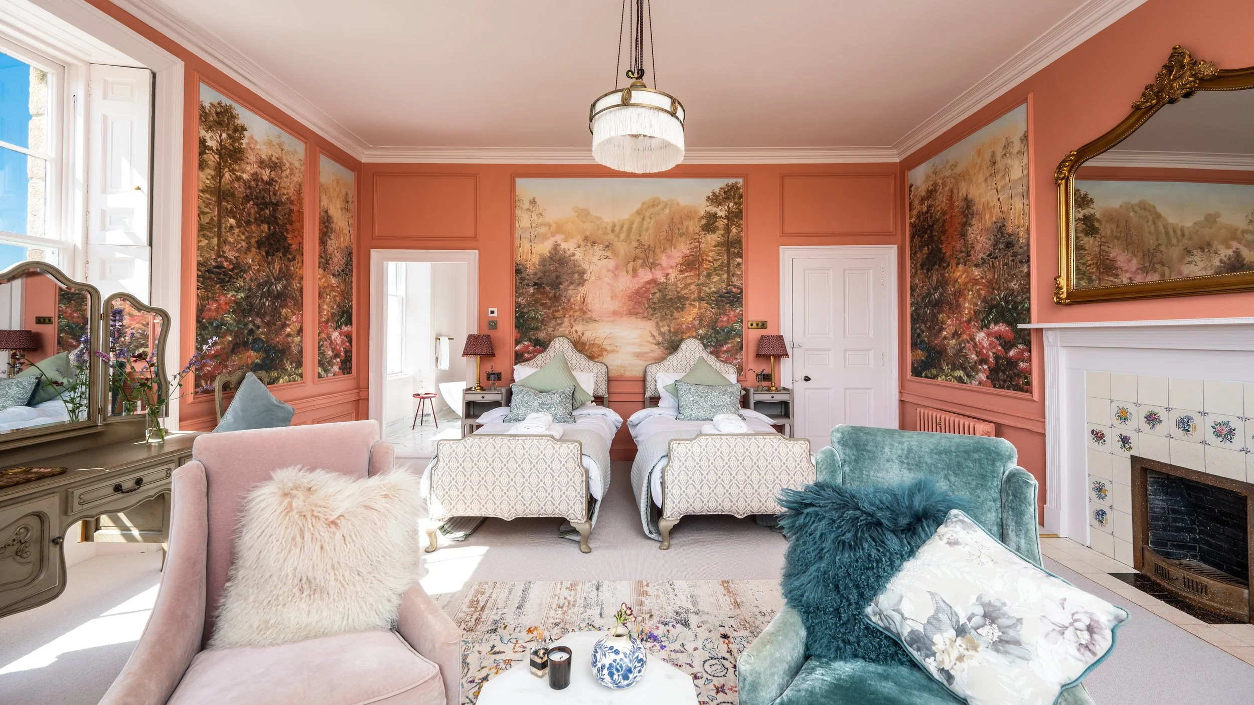 A living room with pink walls, floral landscape murals, a fireplace, and vintage furniture including armchairs with various pillows, a pink sofa, and a large mirror.