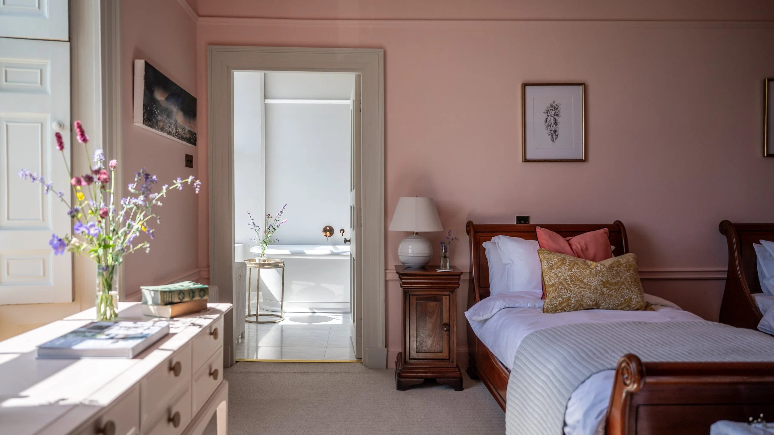 A bedroom with pink walls, a wooden bed with white bedding and gold-patterned pillows, a bedside table with a lamp and a small vase, a dresser with flowers and books, a doorway leading to a bathroom with a bathtub, and framed pictures on the walls.