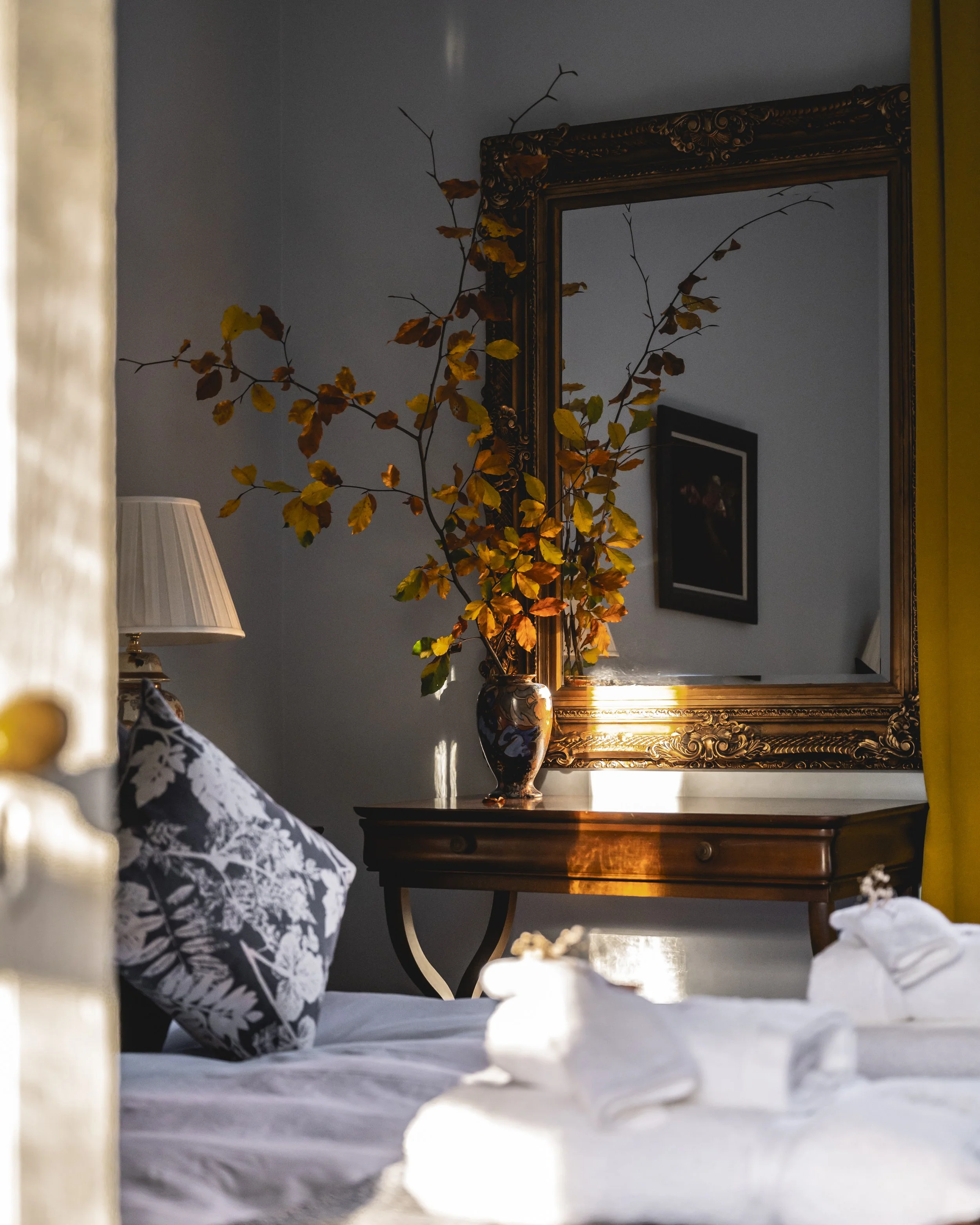 A bedroom with a reflection in a large ornate gold-framed mirror above a wooden table. A decorative vase with branches and yellow leaves, a table lamp with a white shade, and a framed picture on the wall are visible in the reflection. The bed has a black and white floral patterned pillow and white linens.