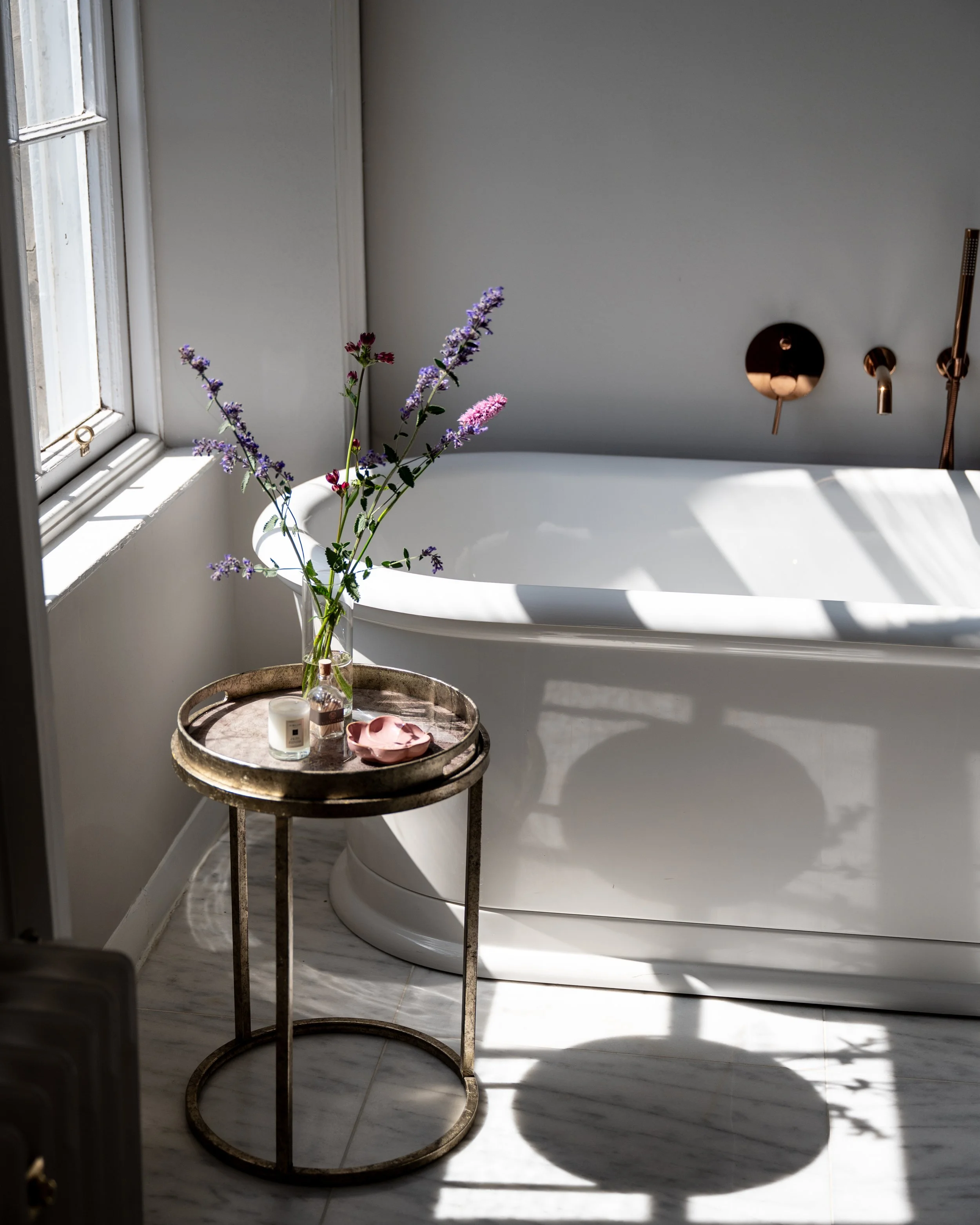 A vase with purple and pink flowers on a small round metal table next to a bathtub in a bathroom, with sunlight casting shadows on the floor.