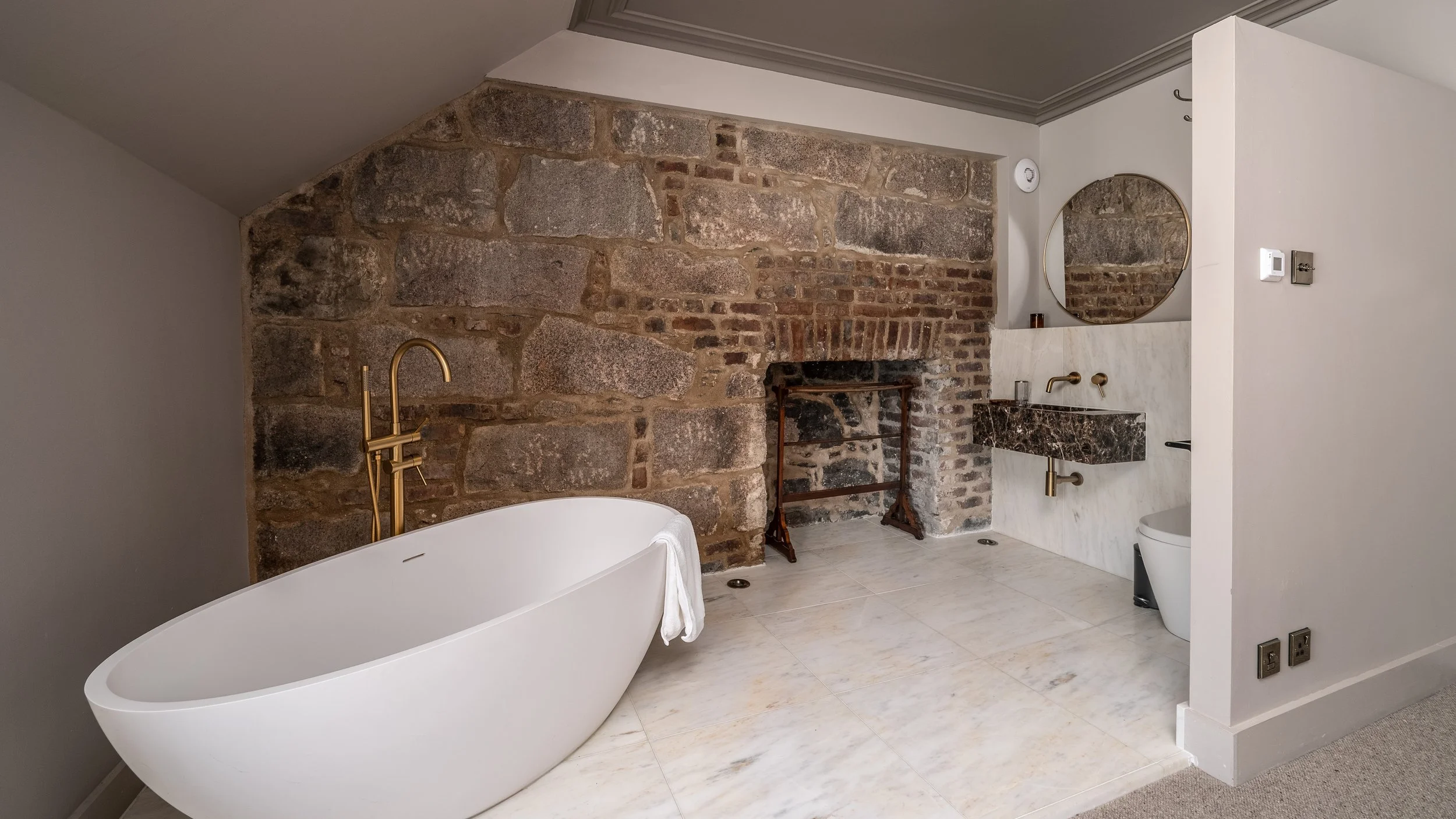 Modern bathroom with a freestanding bathtub, exposed brick and stone wall, gold fixtures, a small marble sink, a round mirror, and some bathroom essentials.